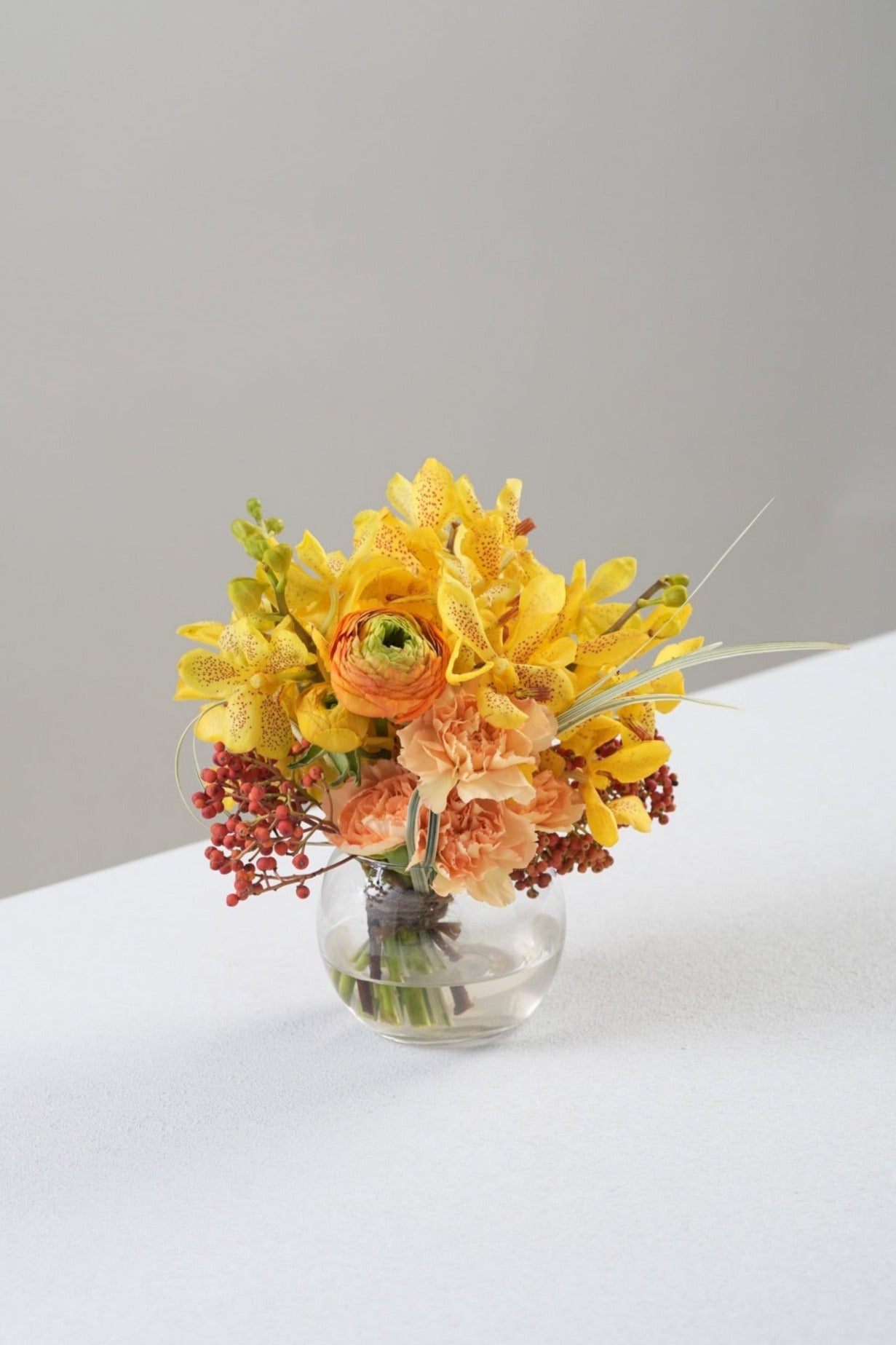 Bring instant cheer with Avah, a mini flower vase arrangement of yellow mokara orchids, orange ranunculus, and red berries. A vibrant floral décor from Just Bloom Hong Kong for desks or gifting.