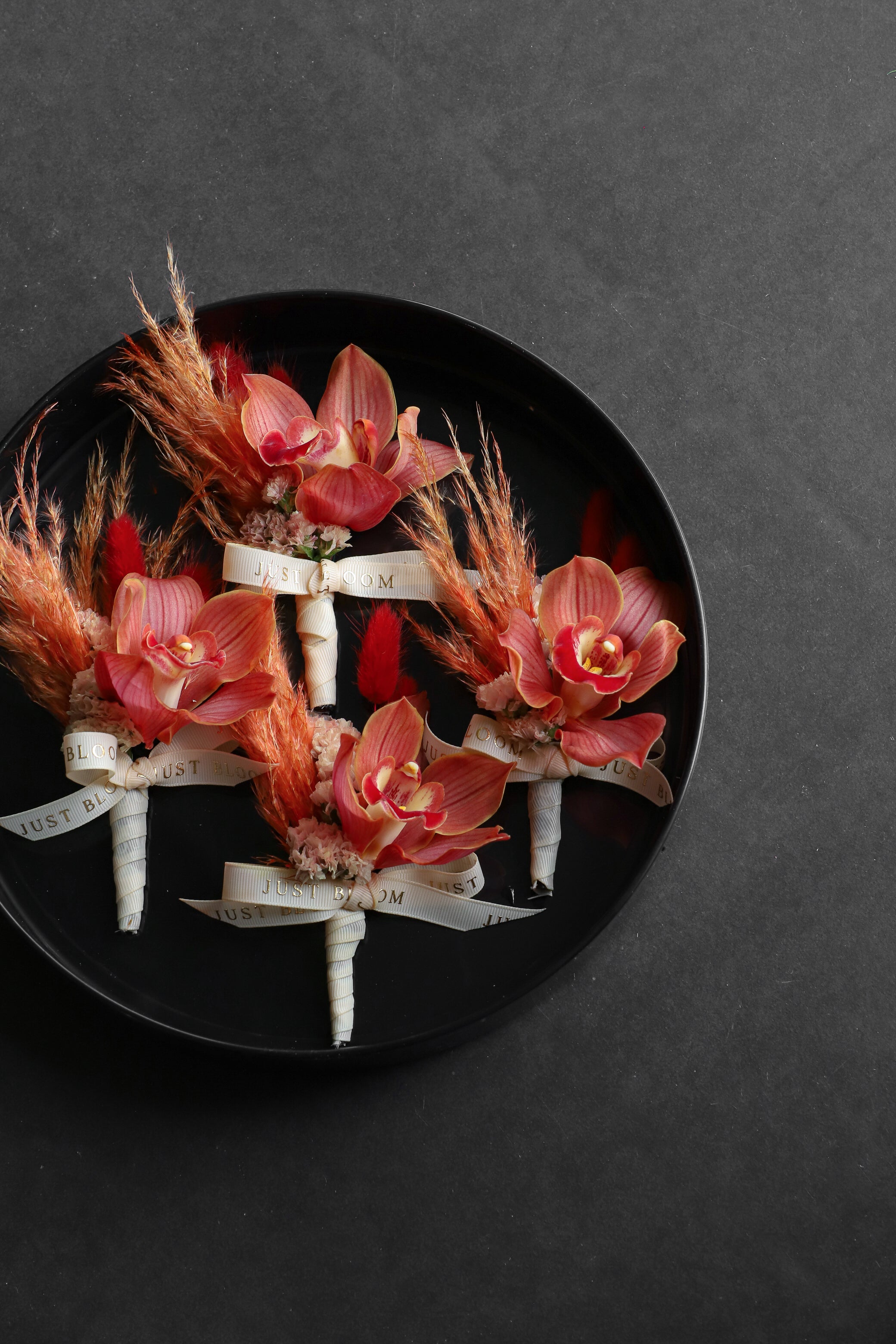 Four Boutonnieres arranged on a black tray featuring cymbidium orchids, myosotis, dried lagurus ovatus and foliage tied with Just Bloom ivory ribbon — Just Bloom Hong Kong