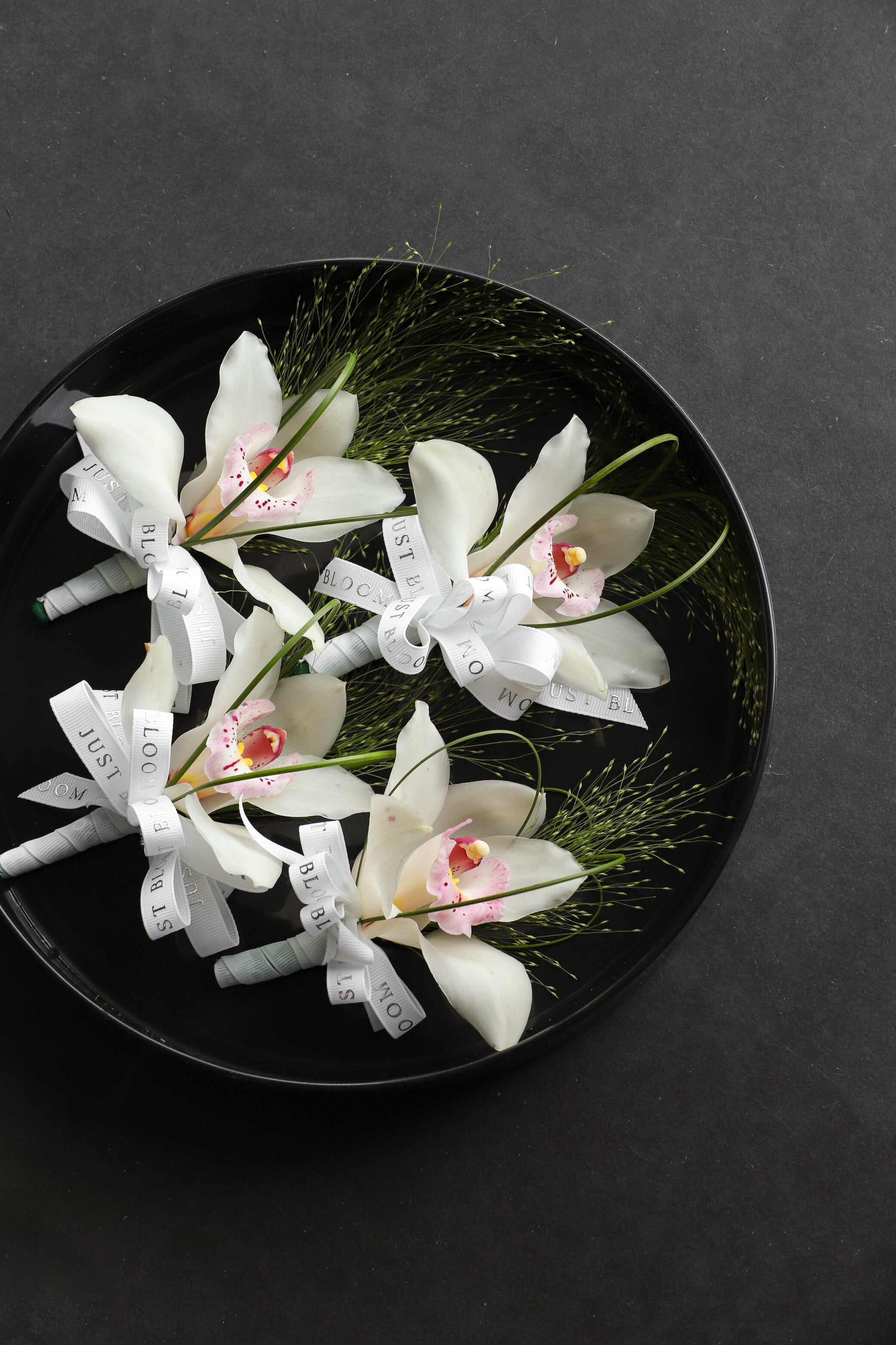 Close-up of Boutonnieres on a black tray by Just Bloom Hong Kong, showcasing white cymbidium orchids with frosted explosion grass, steel grass and Just Bloom white ribbon