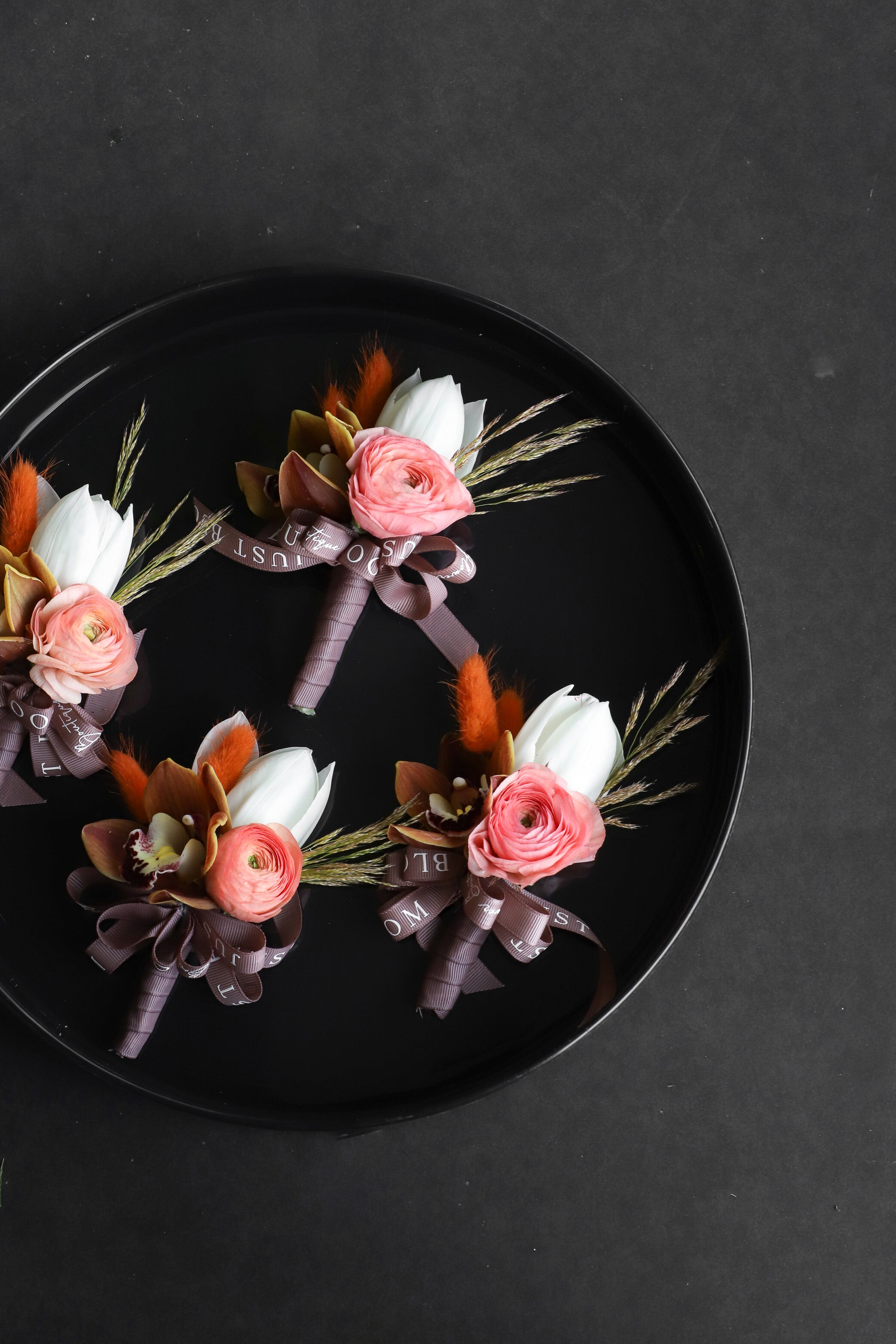 Close-up of four Boutonnieres on a black tray by Just Bloom Hong Kong, showcasing white tulips, coral ranunculus, cymbidium orchids, dried lagurus ovatus and Just Bloom dark ribbon