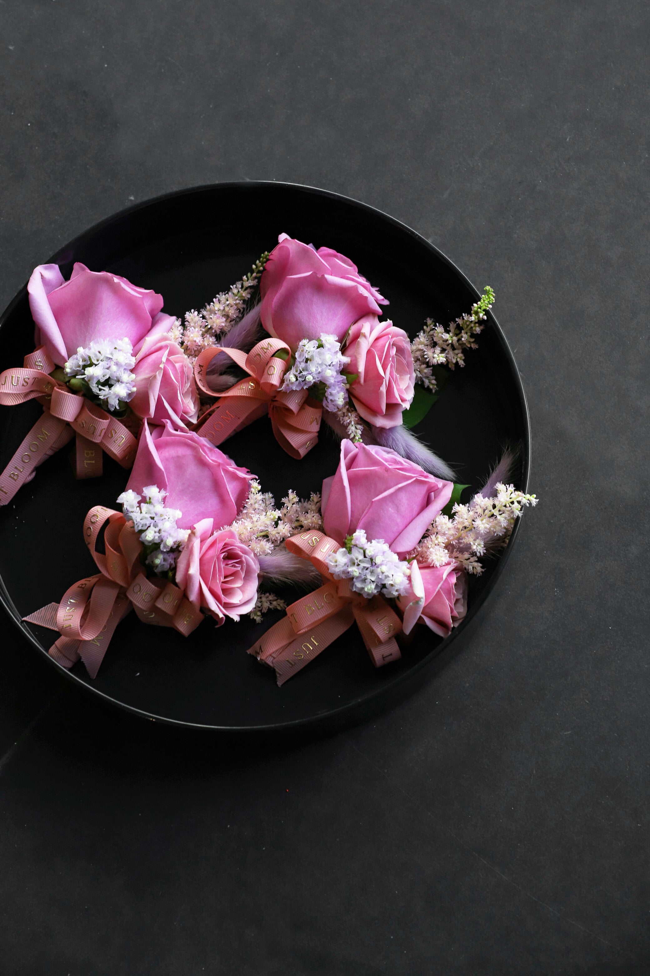 Close-up of four Boutonnieres on a black tray by Just Bloom Hong Kong, showcasing pink roses, pink astilbes, myosotis and feathery accents with Just Bloom rose gold ribbon