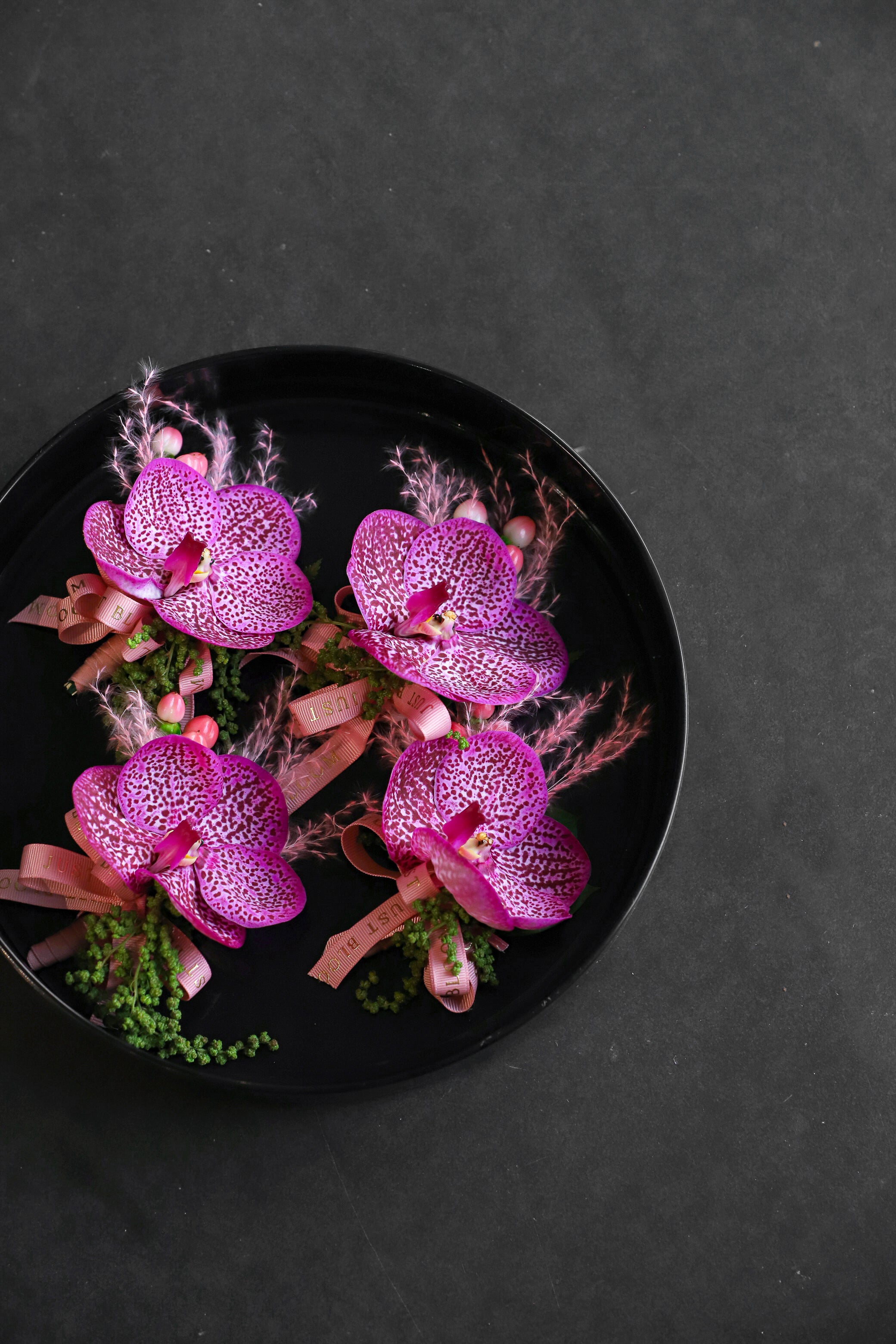 Close-up of four Orchid Edit Boutonnieres on a black tray by Just Bloom Hong Kong, showcasing striking vanda orchids with intricate purple and pink patterning, pink broom bloom and Just Bloom rose gold ribbon