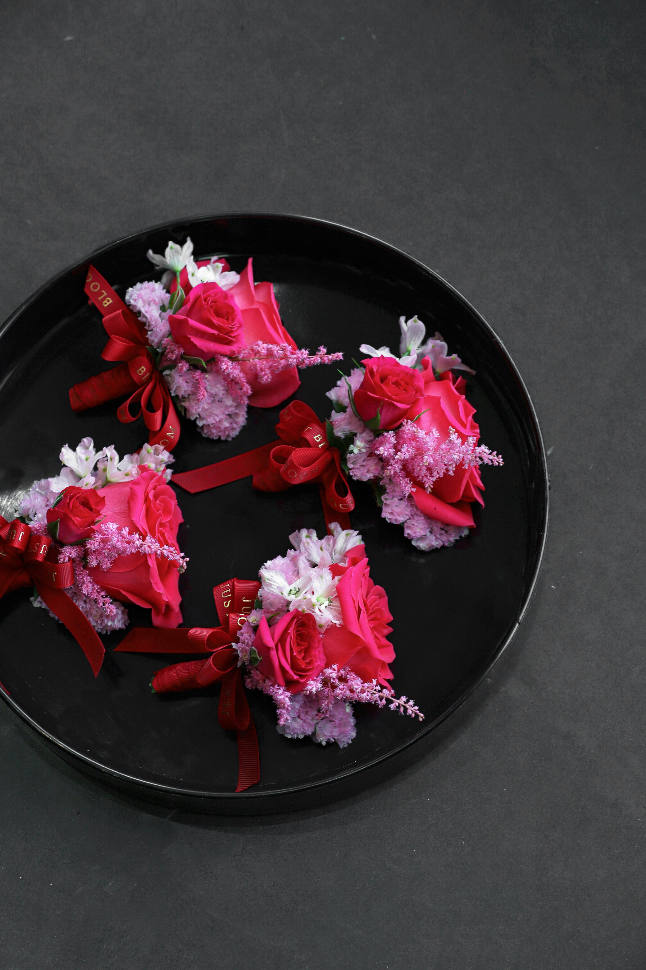 Close-up of four Boutonnieres on a black tray by Just Bloom Hong Kong, showcasing fuchsia roses, pink astilbes, sweet peas and myosotis with Just Bloom deep red ribbon