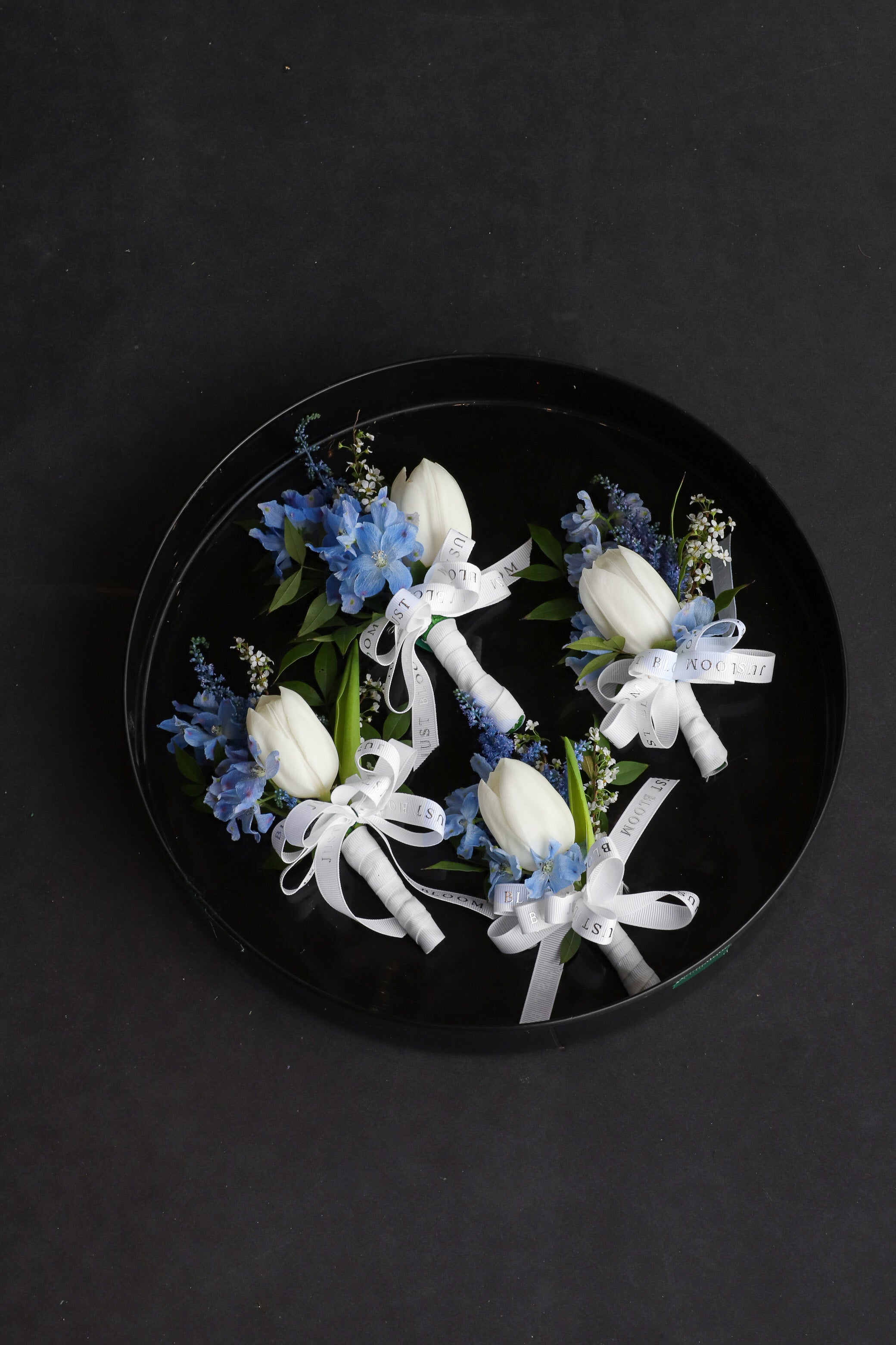 Four Blue Boutonnieres arranged on a black tray featuring white tulips, dusty blue delphiniums and astilbes tied with Just Bloom white ribbon — Just Bloom Hong Kong