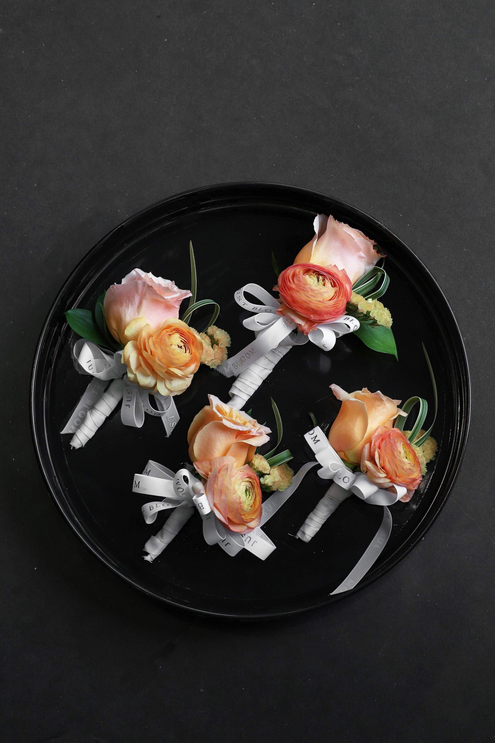 Four Sun Kissed corsages arranged on a black tray featuring peach Ecuadorian roses, orange ranunculus and myosotis tied with Just Bloom white ribbon — Just Bloom Hong Kong