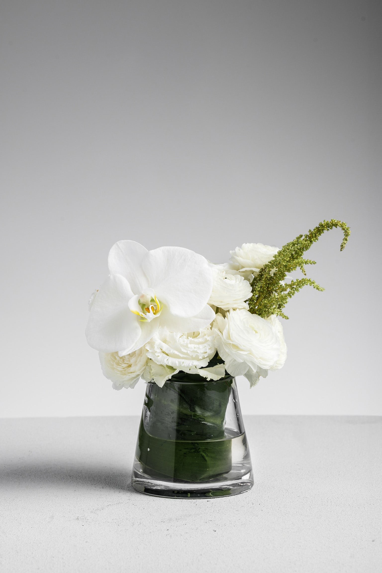 Allyson - A refined mini vase arrangement with white phalaenopsis orchid, ranunculus, and seasonal greens in a clear glass vase. Designed for minimal elegance. Available for same-day delivery in Hong Kong by Just Bloom.