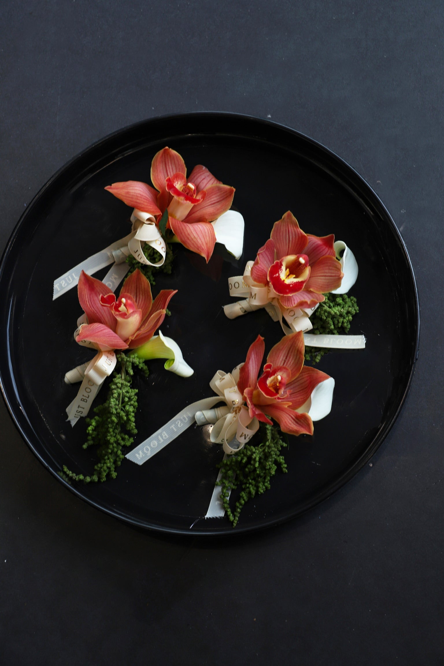 Four Amber Boutonniere arranged on a black tray featuring terracotta cymbidium orchids, white calla lilies and foliage tied with Just Bloom ivory ribbon — Just Bloom Hong Kong