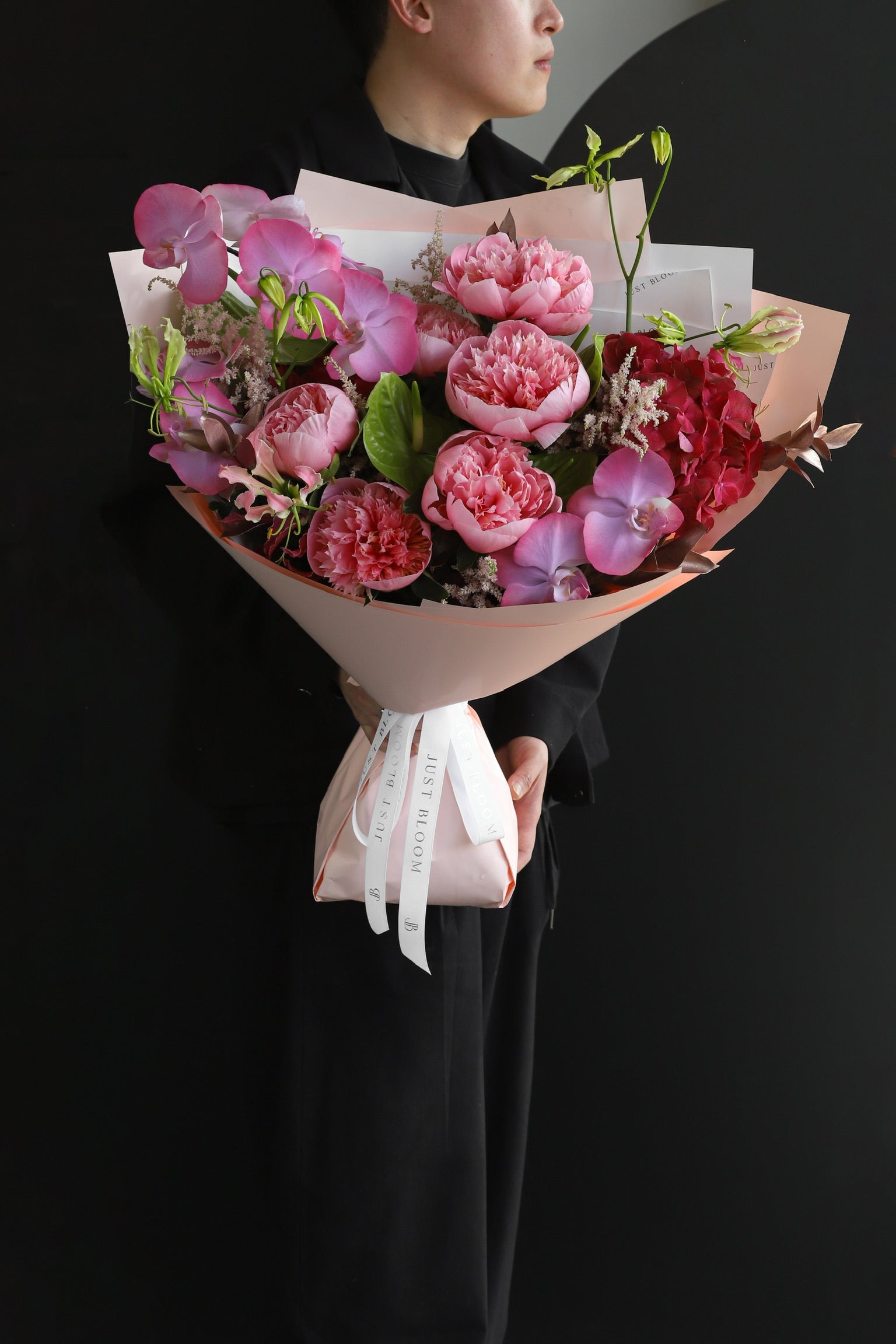 A model in black holding the Bloom Theory luxury bouquet featuring pink peonies, purple phalaenopsis orchids and astilbes wrapped in blush pink with a Just Bloom white ribbon — Just Bloom Hong Kong