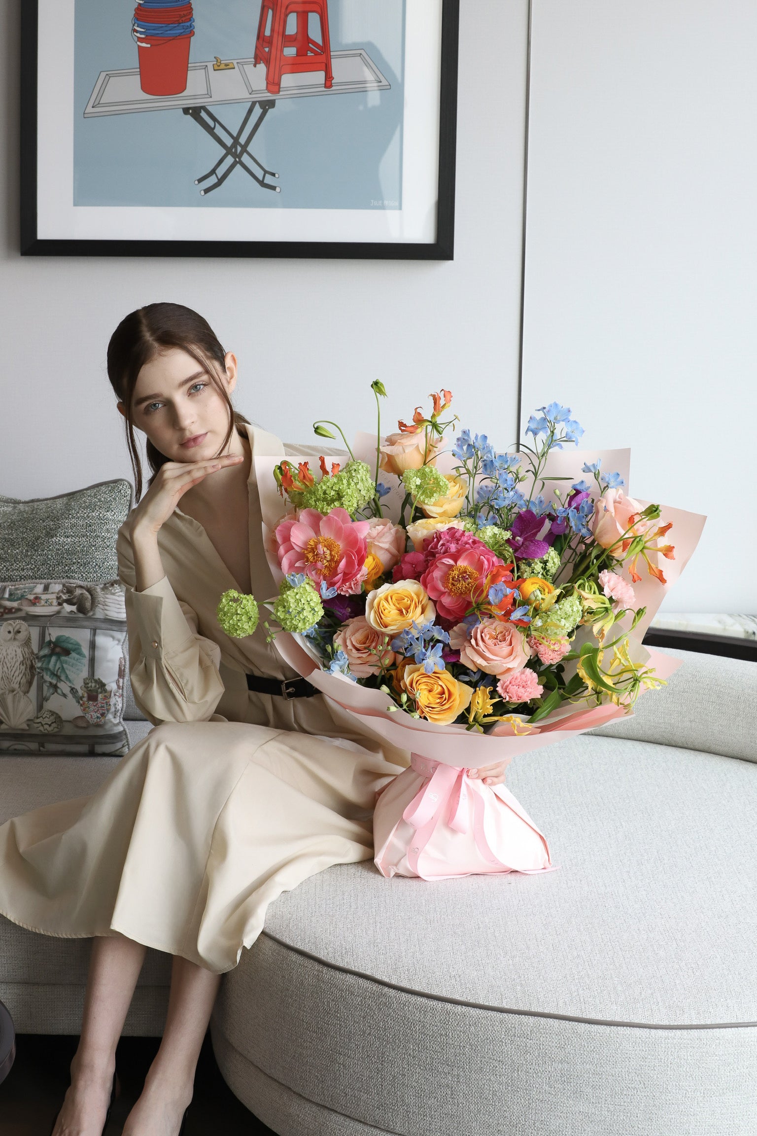 A model seated on a luxury sofa beside Blooming with Love, a large vibrant bouquet in coral, pink, blue and green with pink wrapping and blush ribbon — Just Bloom Hong Kong