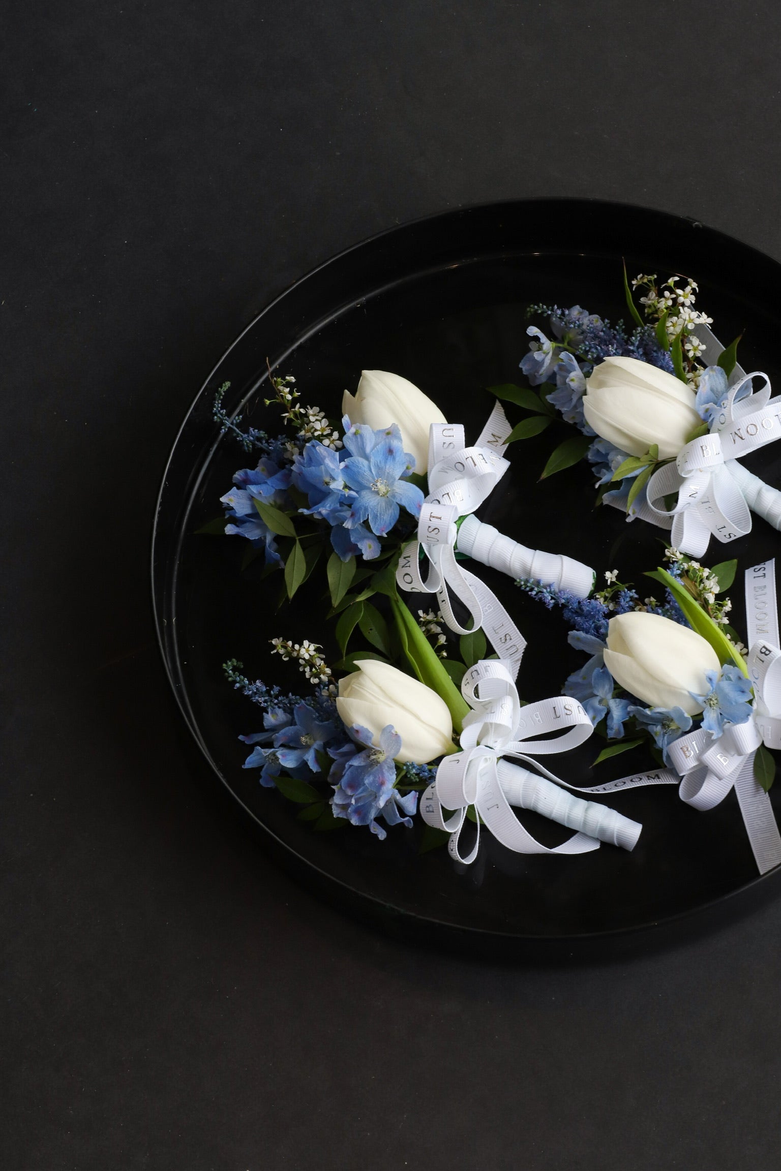 Four Blue Whisper corsages arranged on a black tray featuring white tulips, dusty blue delphiniums and astilbes tied with Just Bloom white ribbon — Just Bloom Hong Kong
