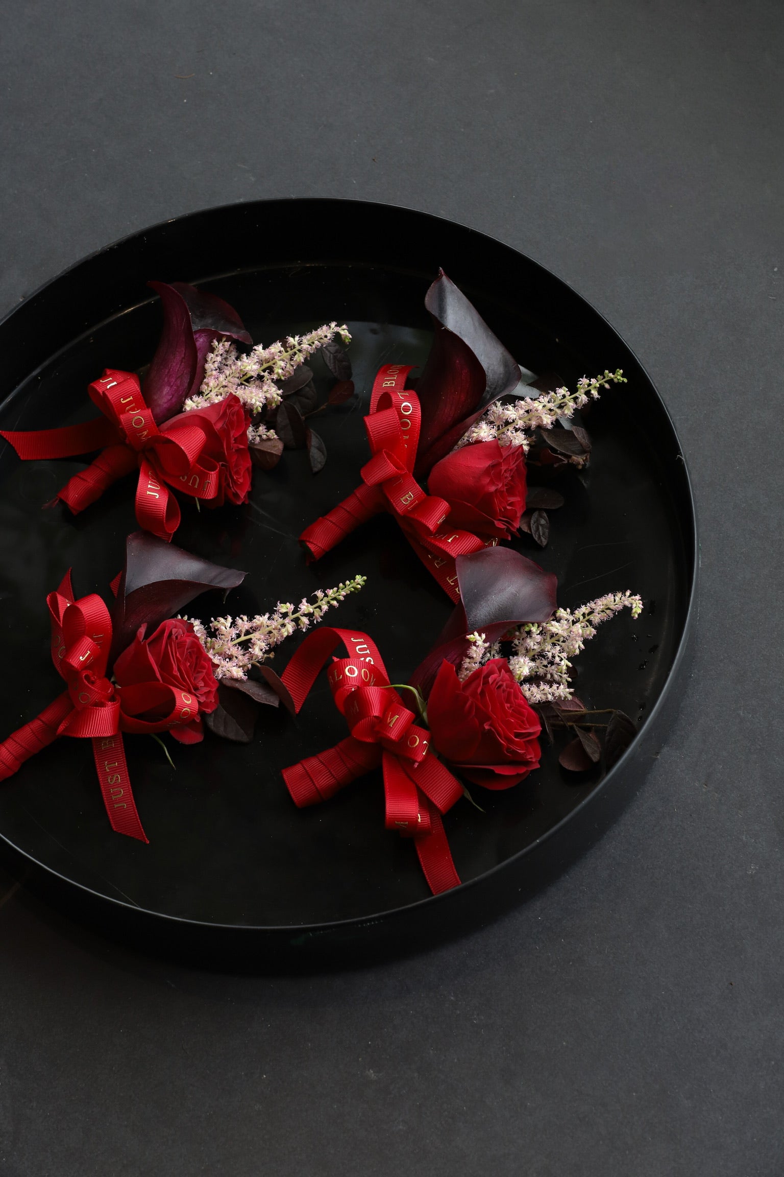 Four Bold Promise corsages arranged on a black tray featuring deep red spray roses, dark calla lilies and astilbes tied with Just Bloom red ribbon — Just Bloom Hong Kong