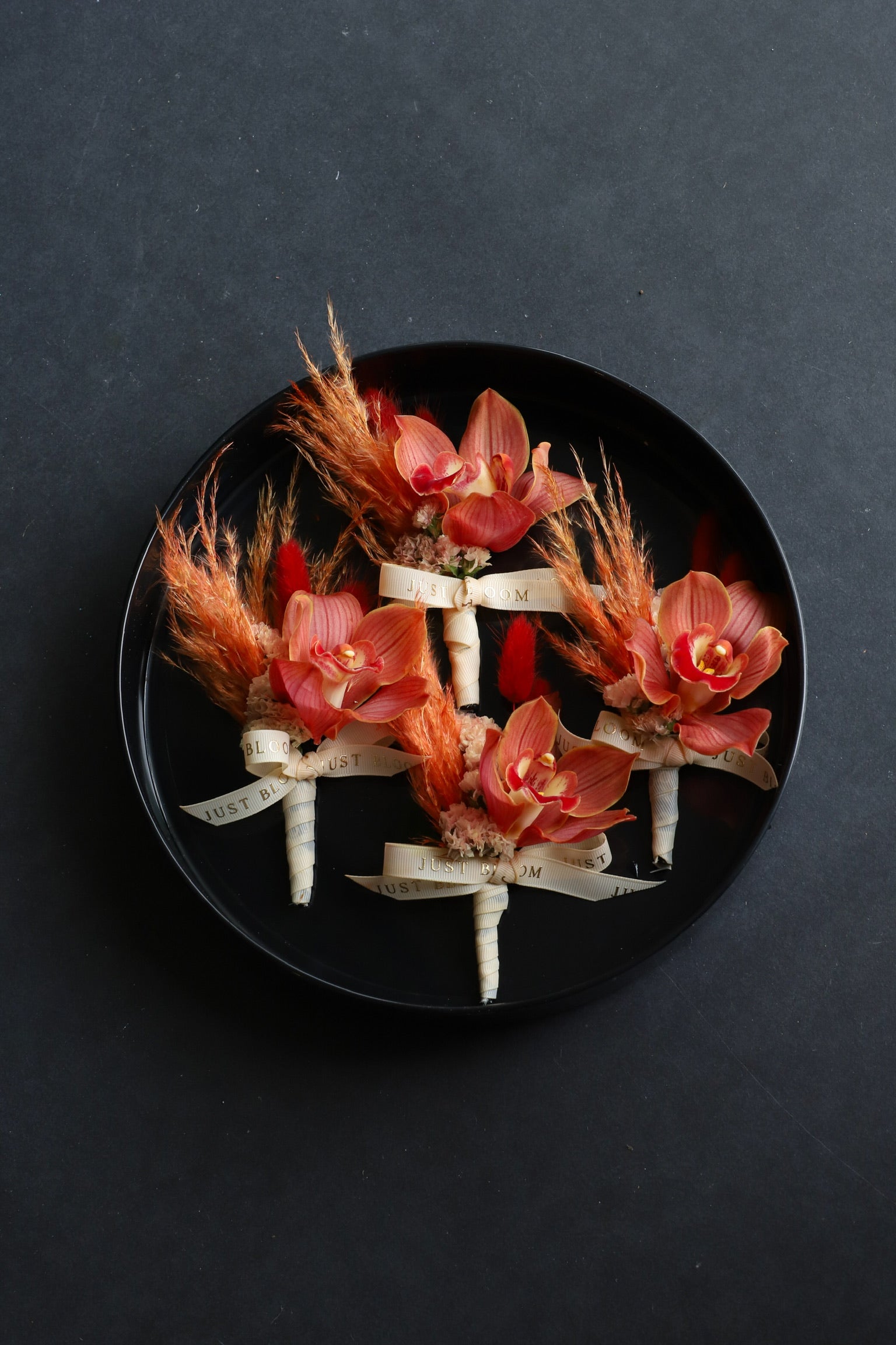 Close-up of four Bound Together wedding corsages on a black tray by Just Bloom Hong Kong, showcasing cymbidium orchids with myosotis, dried lagurus ovatus and Just Bloom ivory ribbon