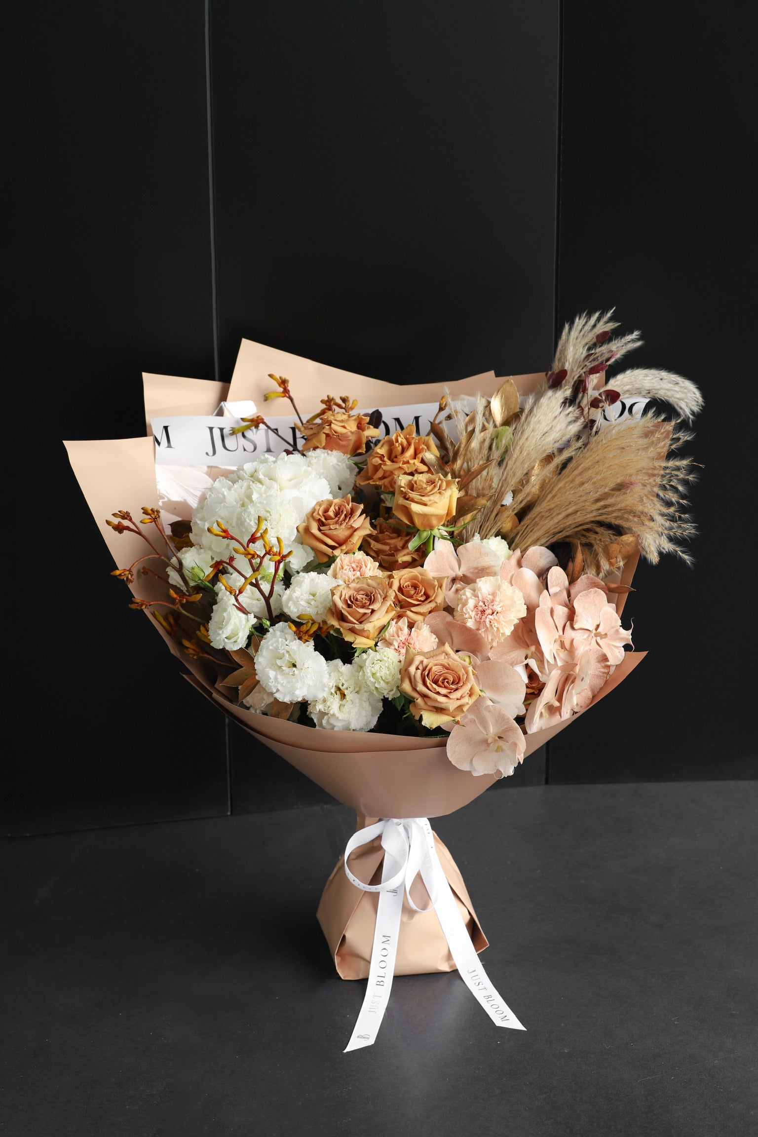 Large warm earth-toned fresh flower bouquet with toffee roses, white blooms, and dried pampas plumes wrapped in nude kraft paper against a dark background — Just Bloom Hong Kong