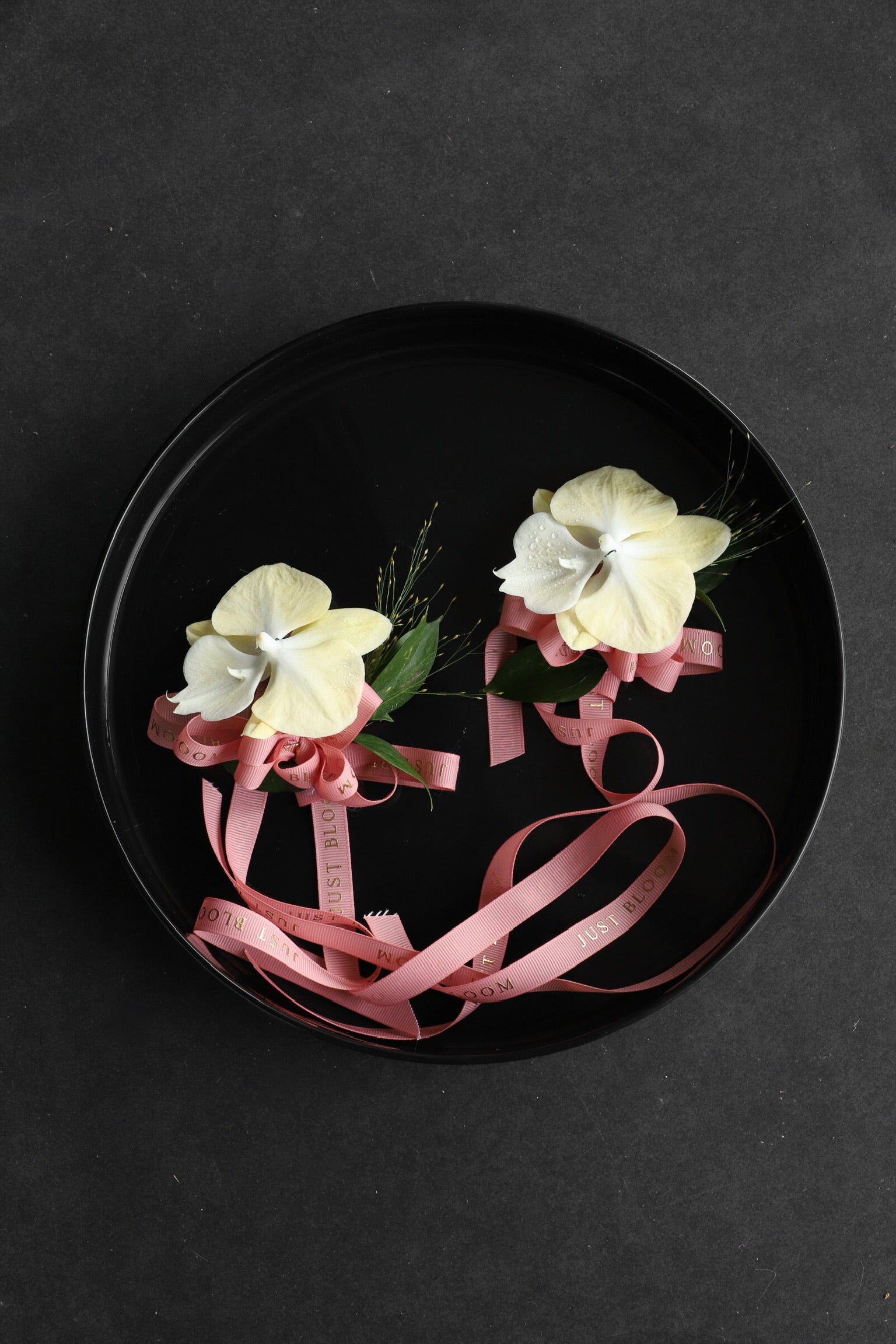 Two Corsage No.2 pieces arranged on a black tray featuring cream phalaenopsis orchids and frosted explosion with Just Bloom pink ribbon — Just Bloom Hong Kong