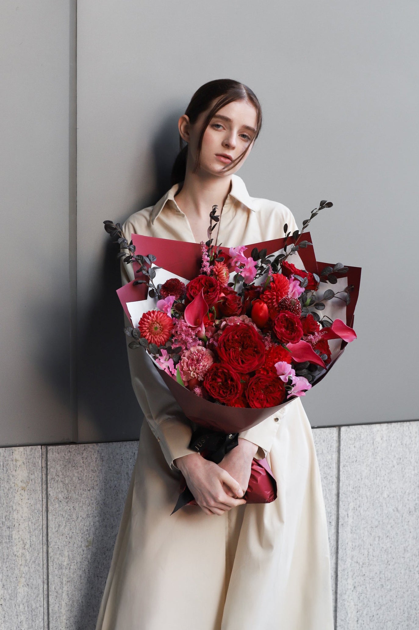 A model leaning against a stone wall holding Deeply Devoted bouquet by Just Bloom Hong Kong, featuring rich crimson and deep red blooms with trailing dark foliage in burgundy wrapping