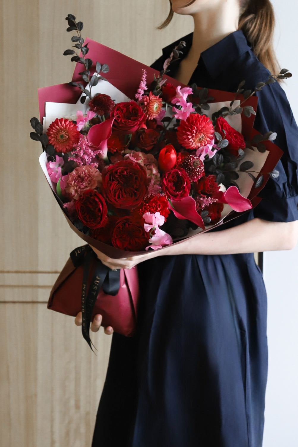A model in a navy dress holding Deeply Devoted luxury bouquet by Just Bloom Hong Kong, showcasing deep red and crimson blooms with dark foliage in burgundy wrapping