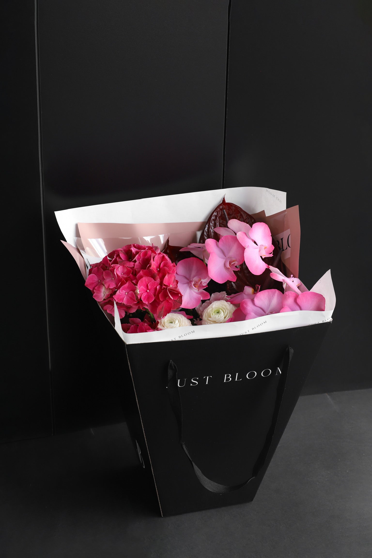 Just Bloom signature black gift bag carrying a large soft pink and peach fresh flower bouquet with mixed blooms and silver foliage against a dark background — Just Bloom Hong Kong