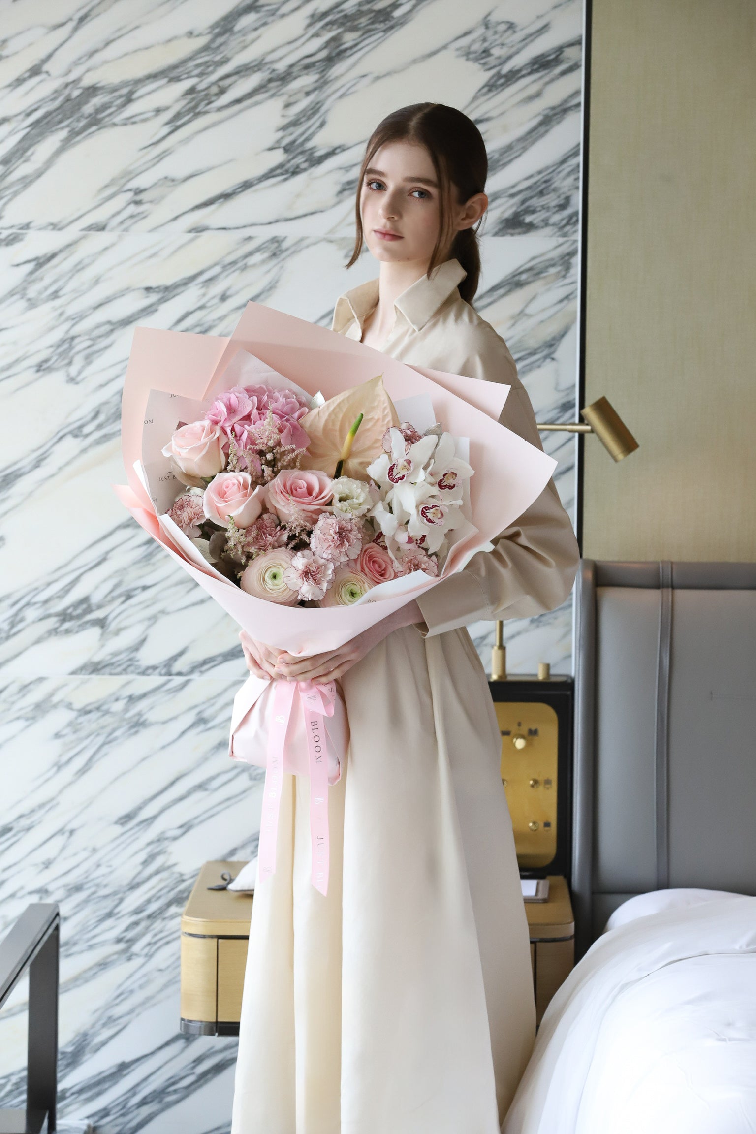 A model standing in a luxury hotel room with marble walls holding Featherlight bouquet by Just Bloom Hong Kong, featuring an exquisitely delicate arrangement of blush and pale pink blooms with pink wrapping and ribbon