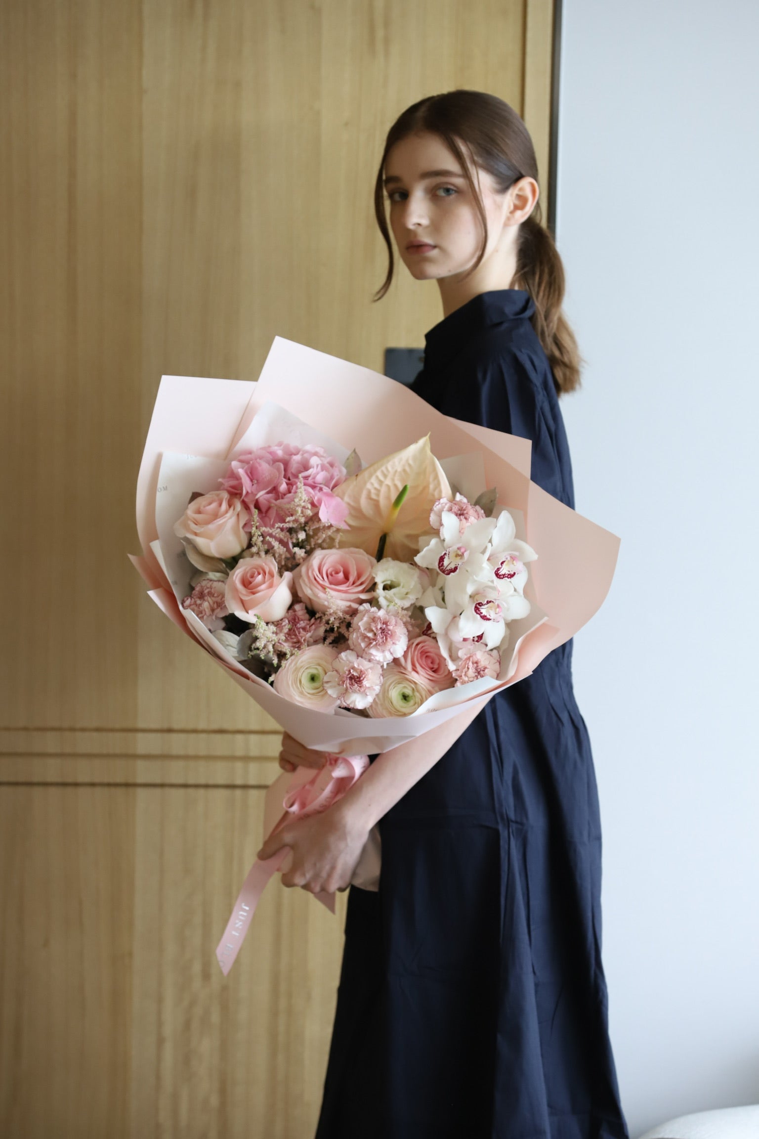 model in a navy dress holding Featherlight luxury bouquet by Just Bloom Hong Kong, showcasing soft blush roses, cymbidium orchids and pale pink blooms in pink wrapping