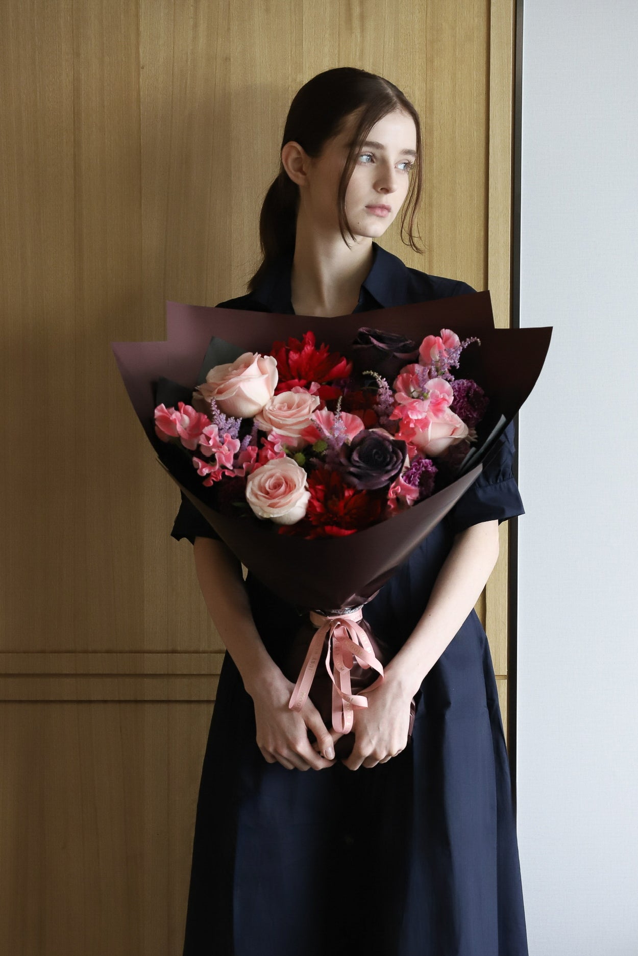 A model in a navy dress holding a Fierce in Bloom luxury Mother's Day bouquet featuring blush roses, deep crimson peonies and dark purple roses, wrapped in burgundy with a pink ribbon — Just Bloom Hong Kong