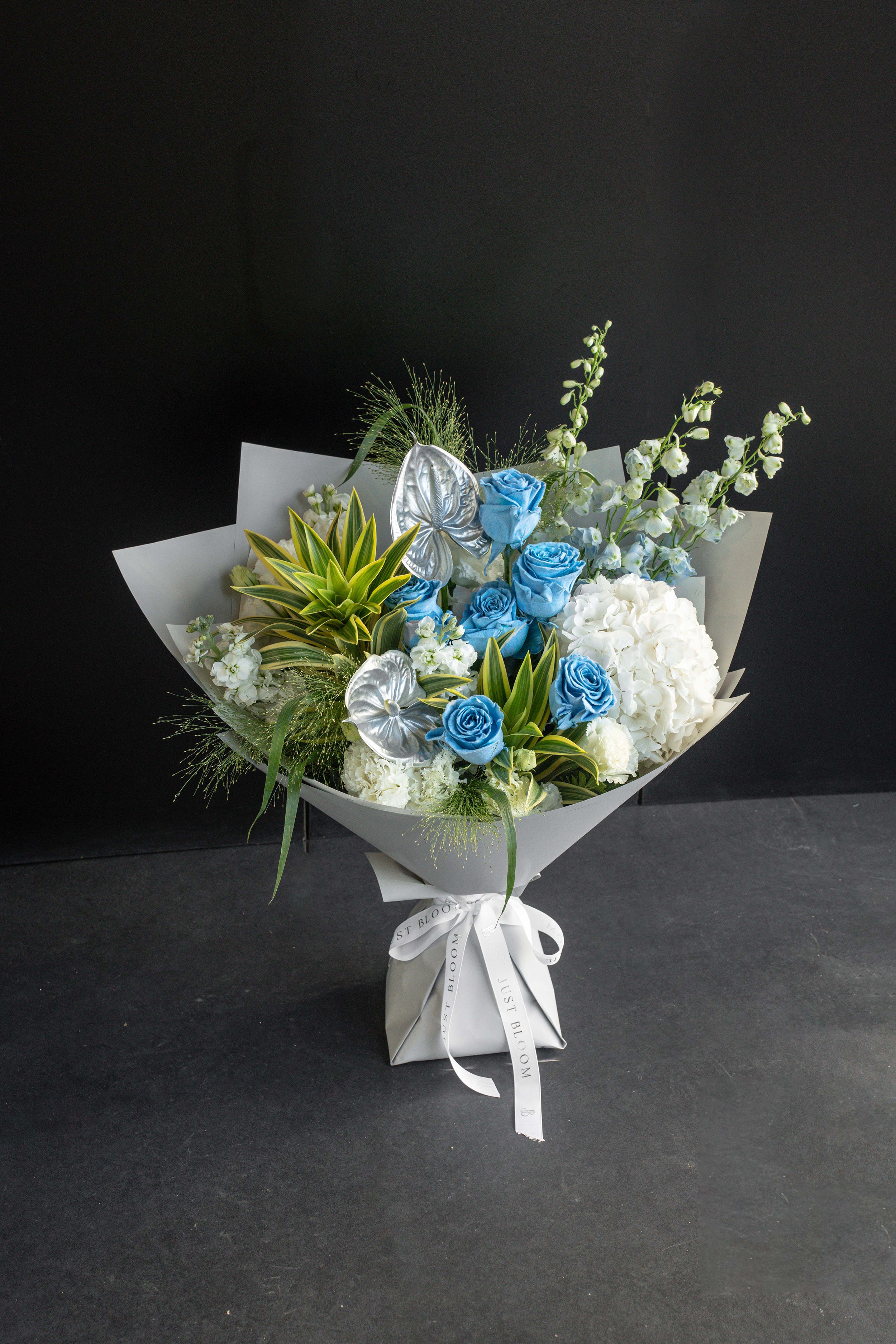 Frost Bloom bouquet of blue roses, white hydrangea, and silver anthurium wrapped in grey paper against a black backdrop — Just Bloom Hong Kong