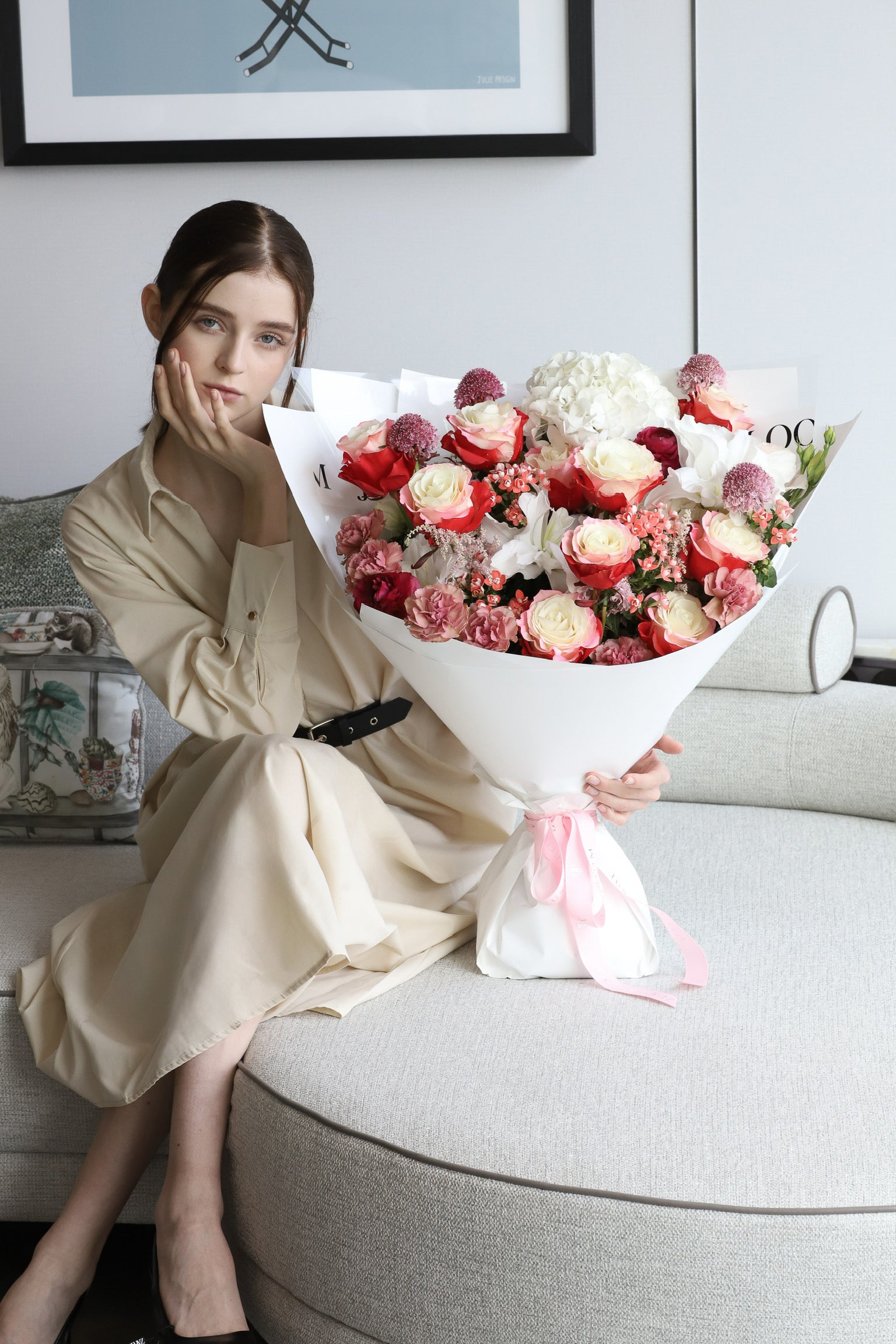 A model seated on a luxury sofa holding Given with Love, a large bouquet in coral, blush and ivory with white wrapping and pink ribbon — Just Bloom Hong Kong