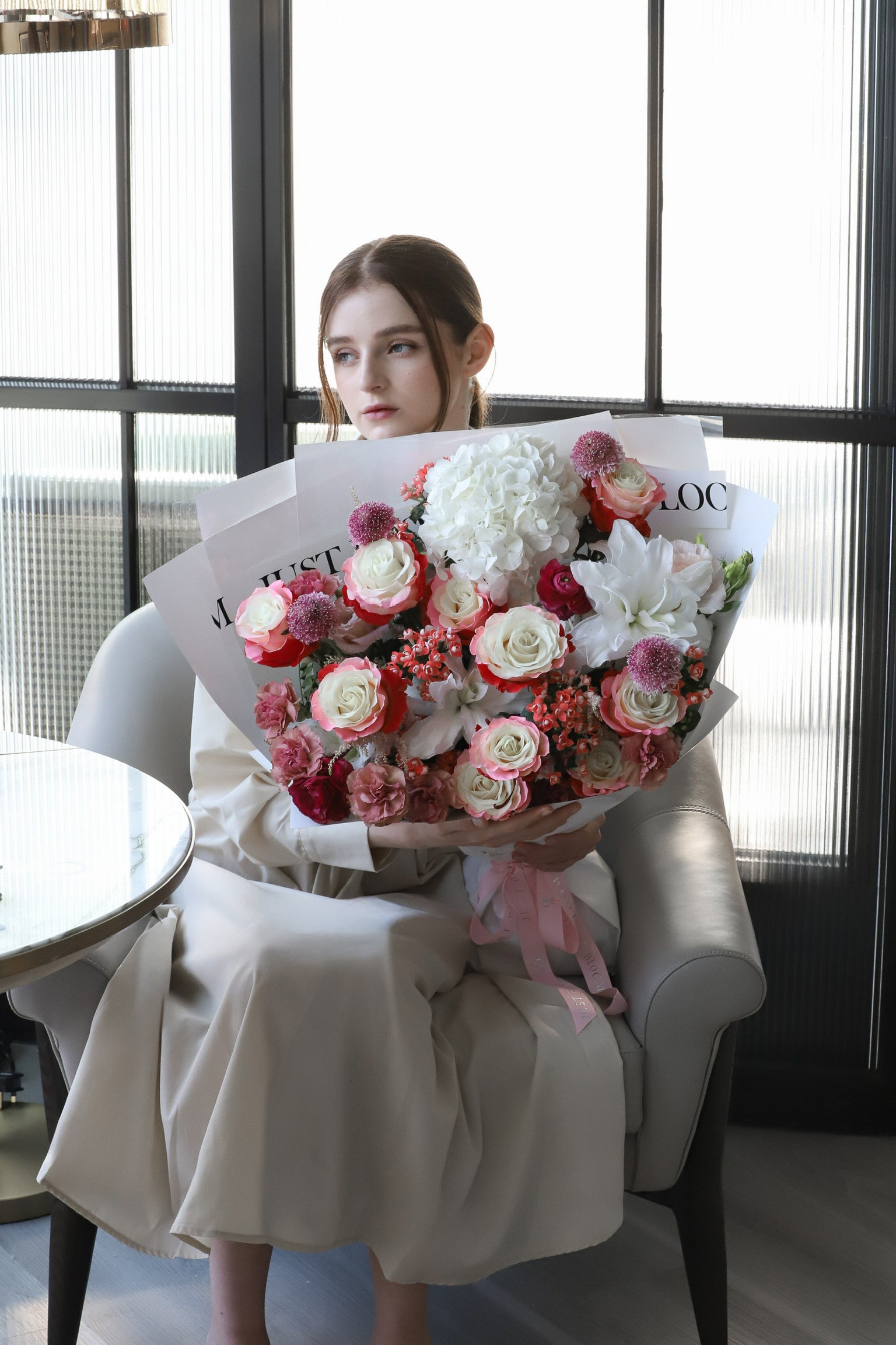 A model on a luxury sofa presenting Given with Love, a generous full-bodied bouquet in warm coral and ivory tones, wrapped in white with a pink ribbon — Just Bloom Hong Kong