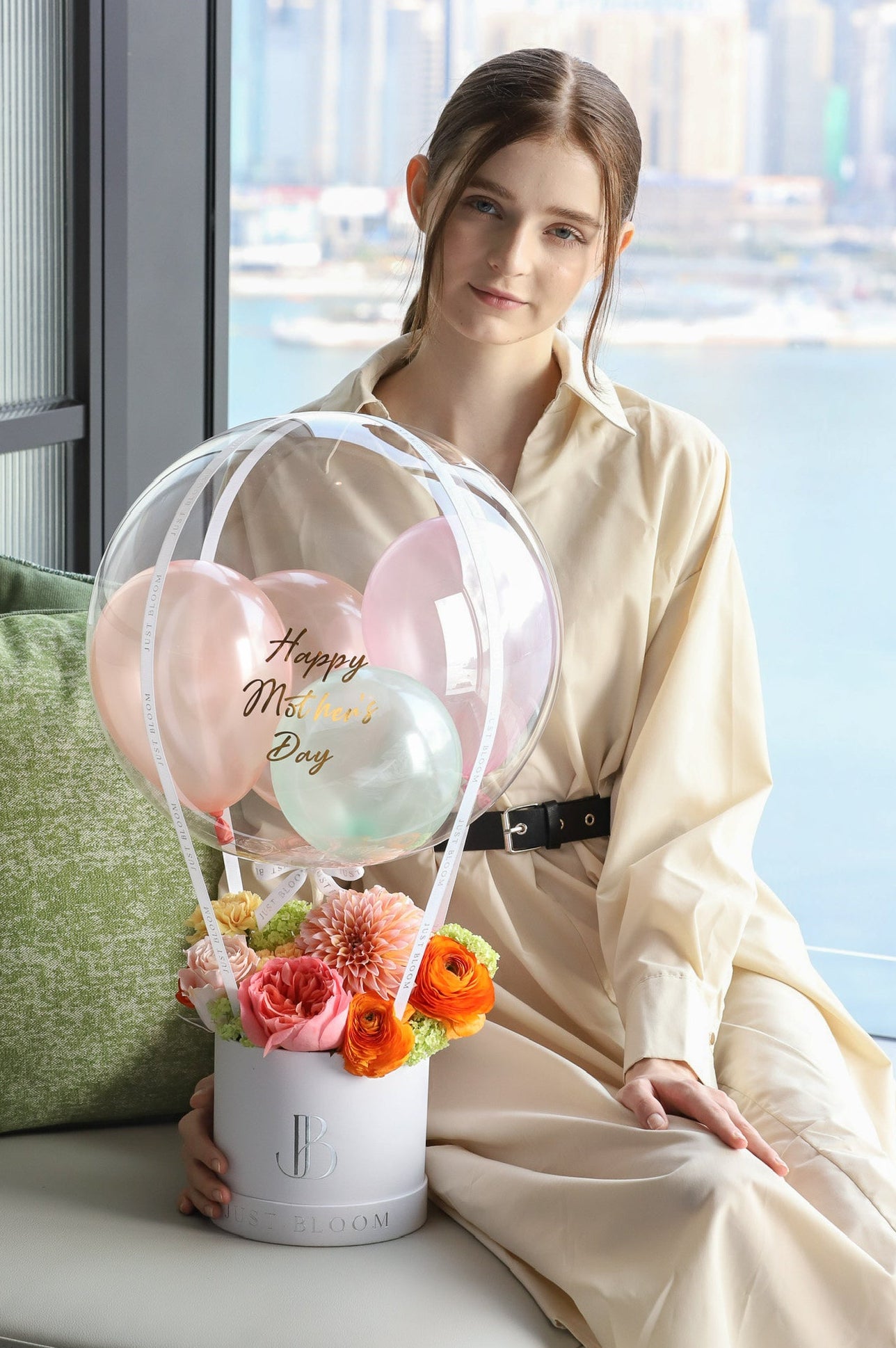 A model holding the Joyful Blooms luxury balloon flower box by Just Bloom Hong Kong with Hong Kong harbour views in the background, showcasing vibrant coral and orange blooms with a personalised balloon arrangement