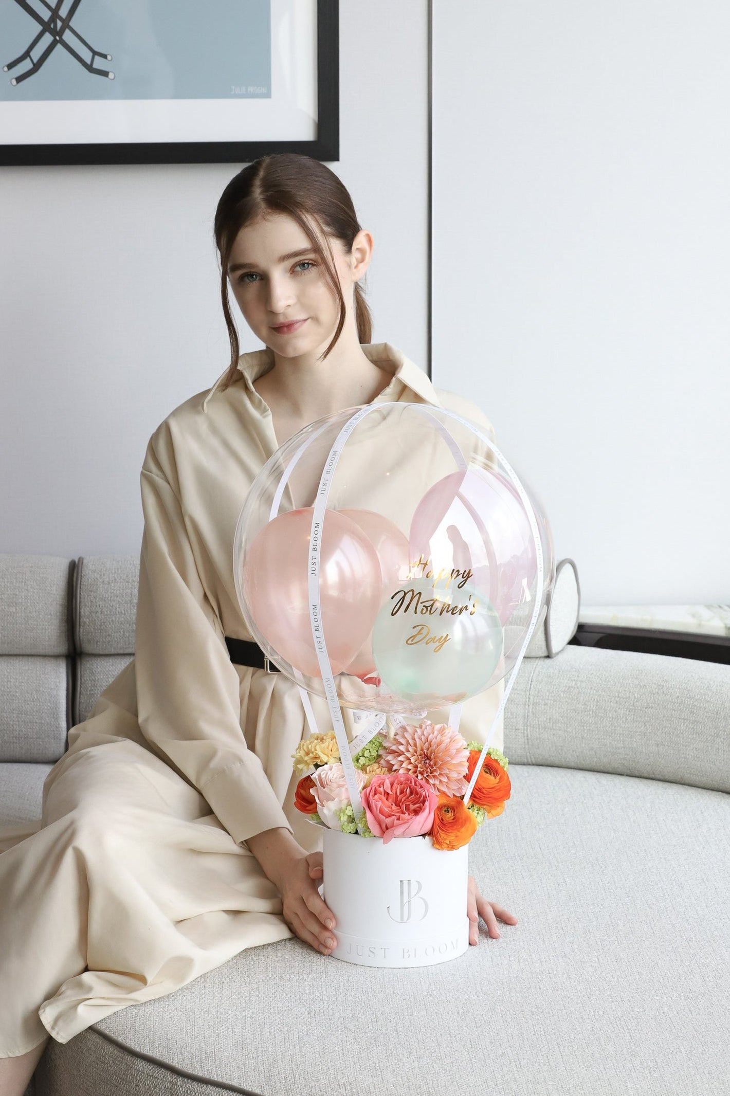 A model seated on a luxury sofa with the Joyful Blooms balloon flower box by Just Bloom Hong Kong, featuring coral and peach blooms in a white box topped with a transparent bubble balloon with Happy Mother's Day gold script