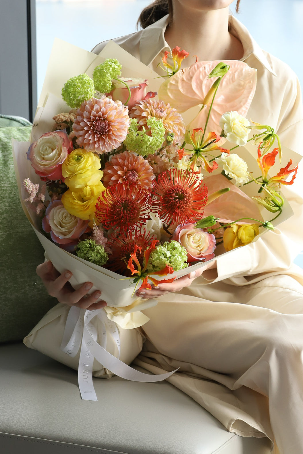 A model seated by a harbour view window holding Luminous, a striking bouquet in warm orange, coral and fresh green with ivory wrapping and white ribbon — Just Bloom Hong Kong