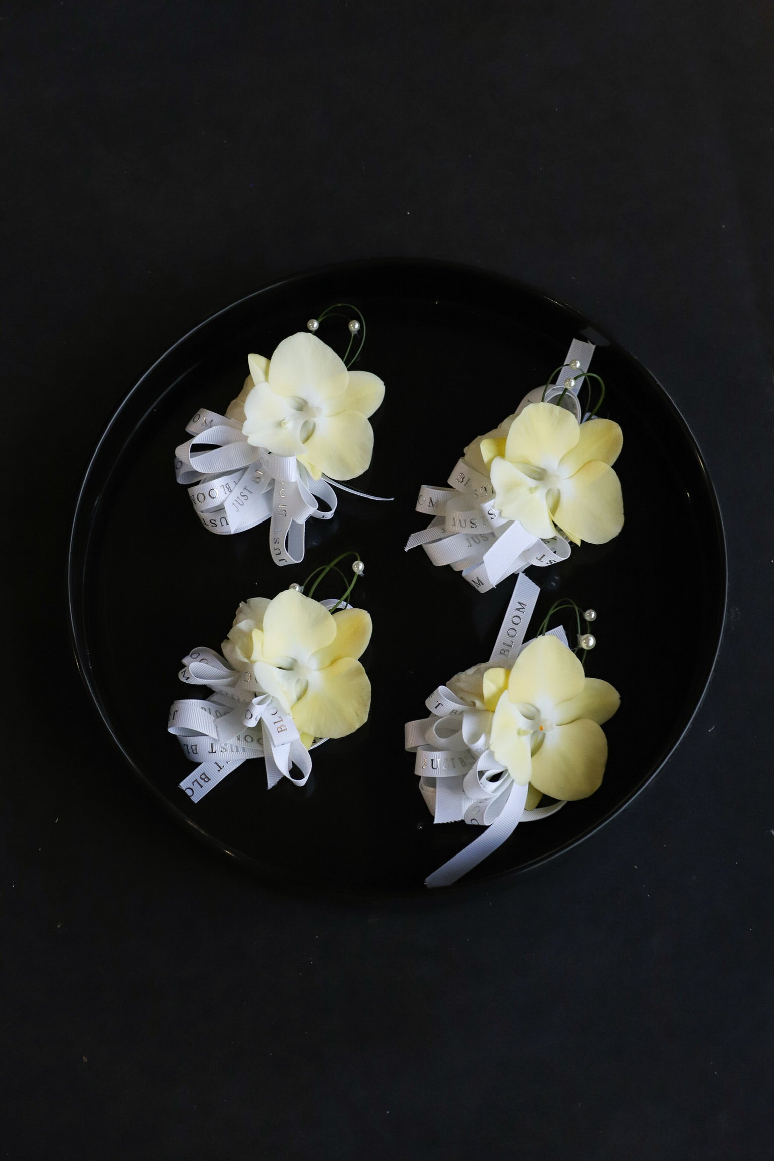 Four Meant to Be corsages arranged on a black tray featuring cream phalaenopsis orchids, steel grass and pearl accents tied with Just Bloom white ribbon — Just Bloom Hong Kong