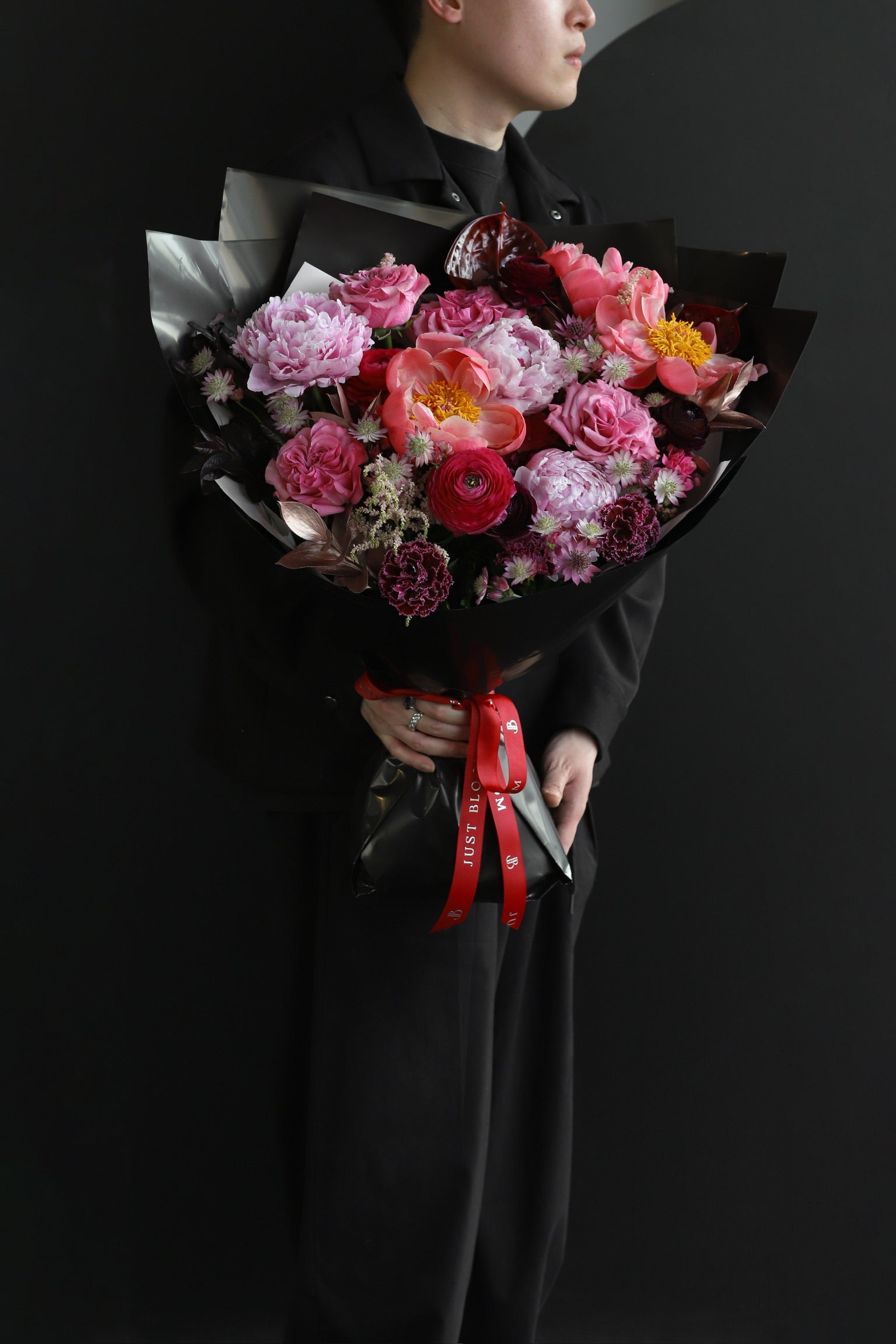 A model in black holding the Midnight Peonies luxury bouquet featuring deep pink peonies, coral ranunculus, roses and burgundy carnations wrapped in black with a Just Bloom red ribbon — Just Bloom Hong Kong