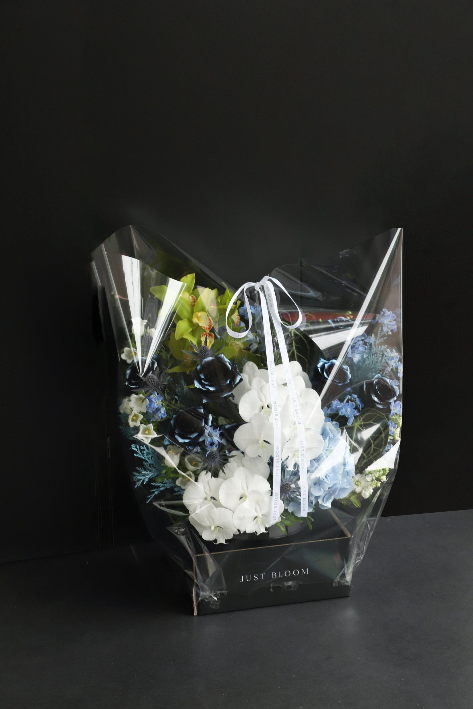 Just Bloom flower box arrangement in clear gift packaging with white ribbon, showing deep navy and white blooms with green Cymbidium Orchids against a dark background — Just Bloom Hong Kong