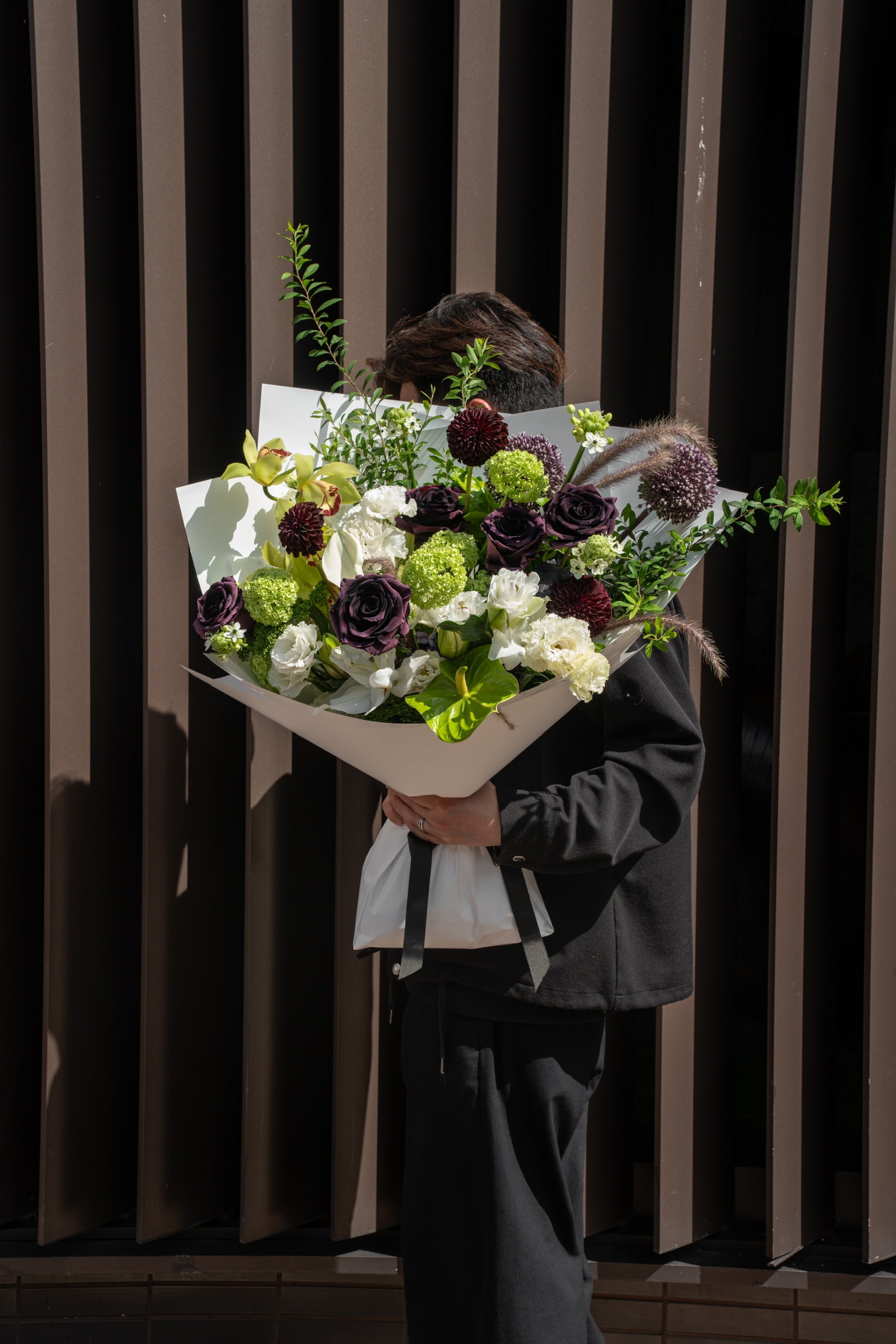 Model holding a large dramatic fresh flower bouquet in deep plum, lime green, and white wrapped in white Just Bloom paper against a dark background — Just Bloom Hong Kong