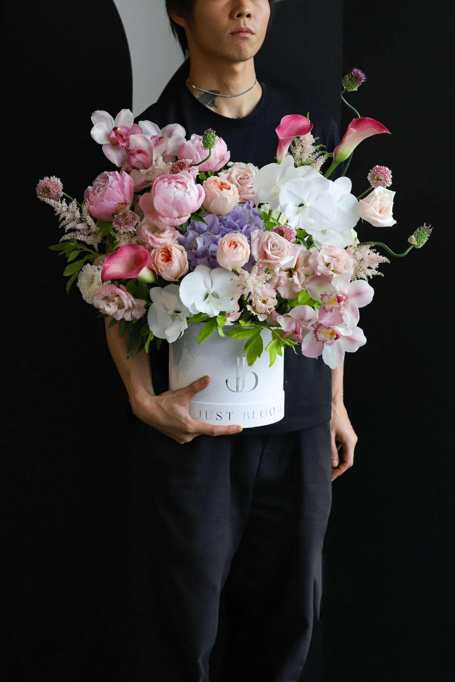 A model in black holding the Opulence luxury flower box by Just Bloom Hong Kong, showcasing an abundant arrangement of peonies, orchids, roses, calla lilies and hydrangeas in a white Just Bloom hat box