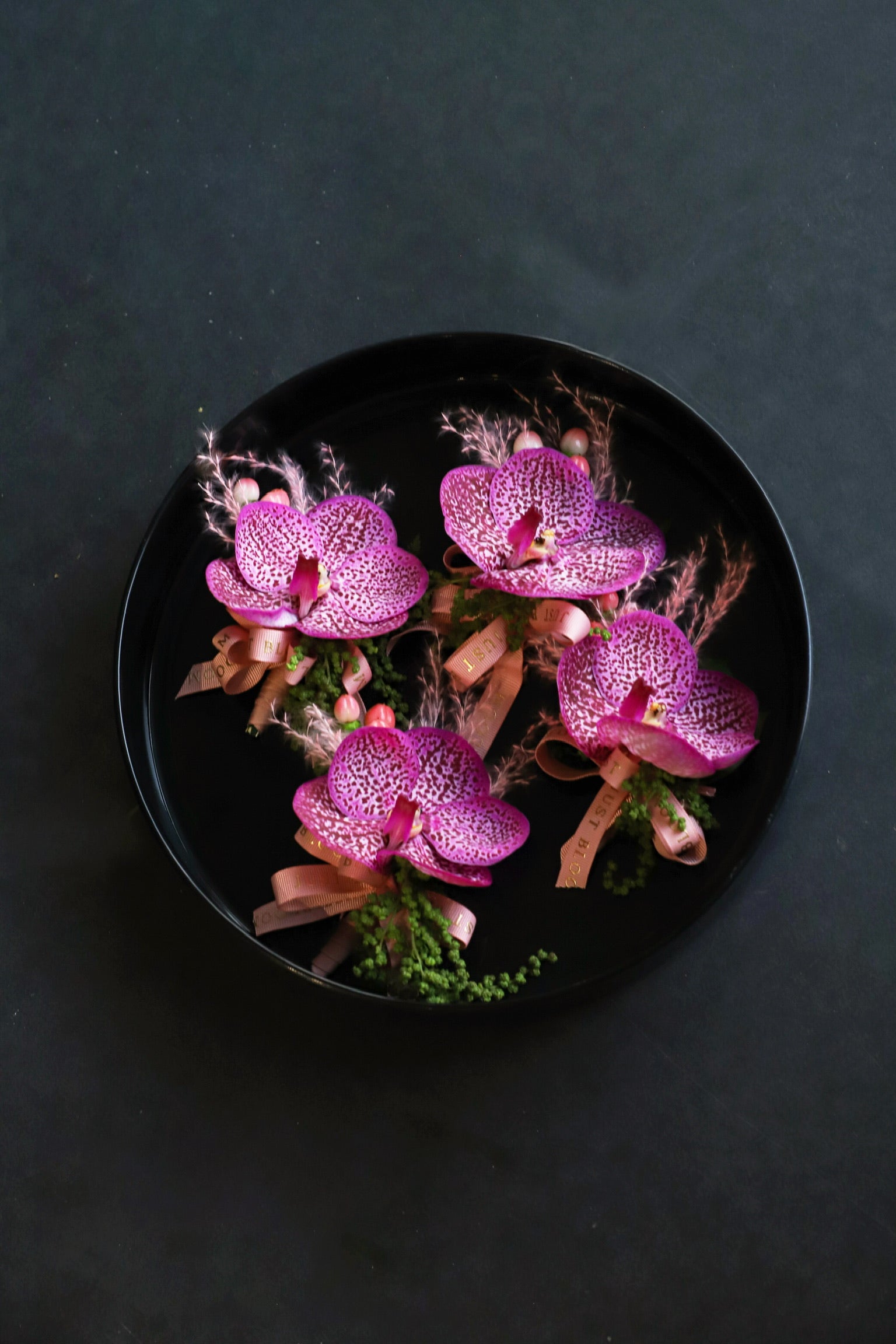 Four Boutonnieres Edit corsages arranged on a black tray featuring purple and pink vanda orchids, pink broom bloom and foliage tied with Just Bloom rose gold ribbon — Just Bloom Hong Kong
