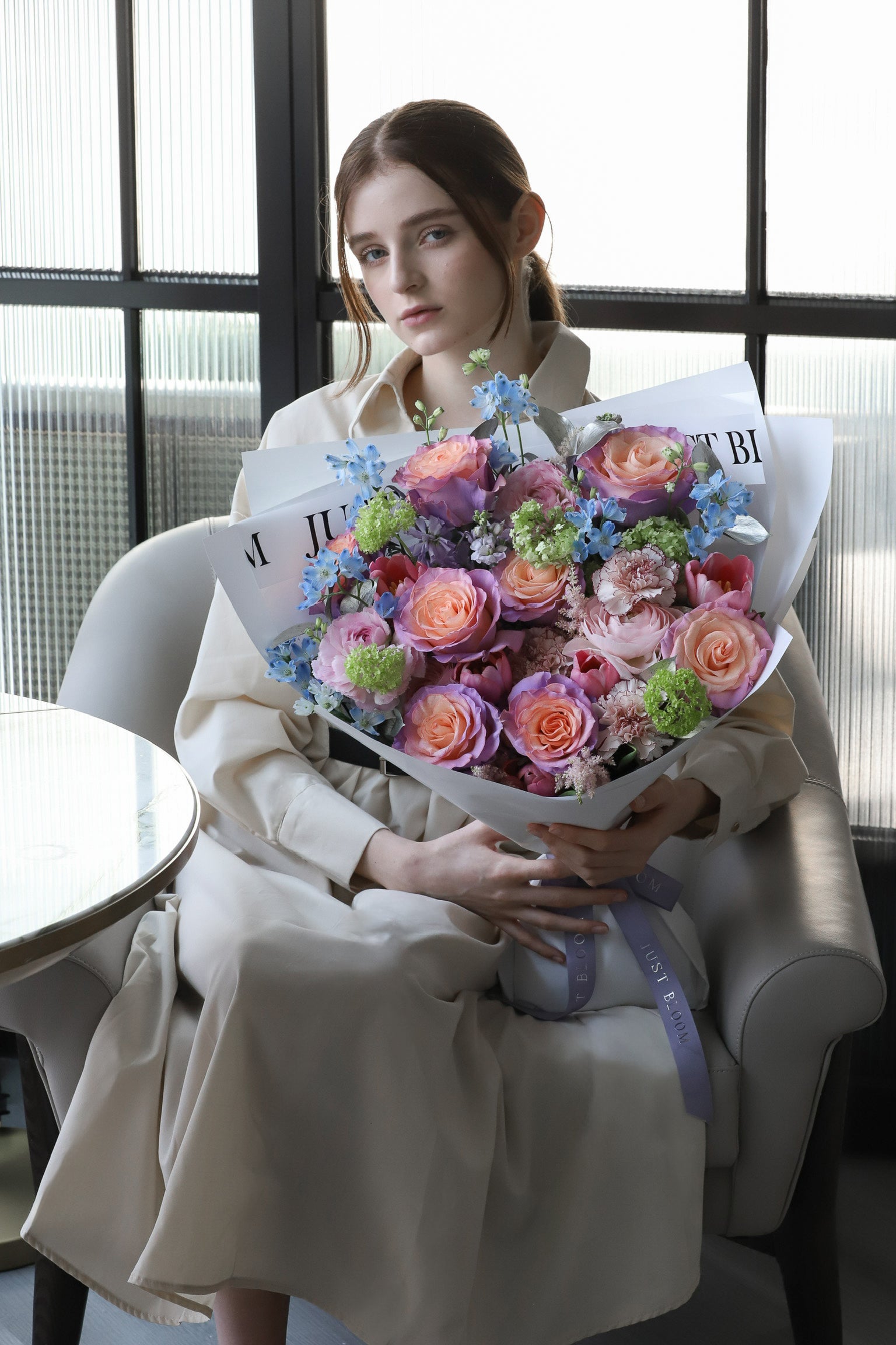 A model seated on a luxury sofa holding Pastel Dreams, a bouquet in soft peach, lavender and blue with white wrapping and lavender ribbon — Just Bloom Hong Kong
