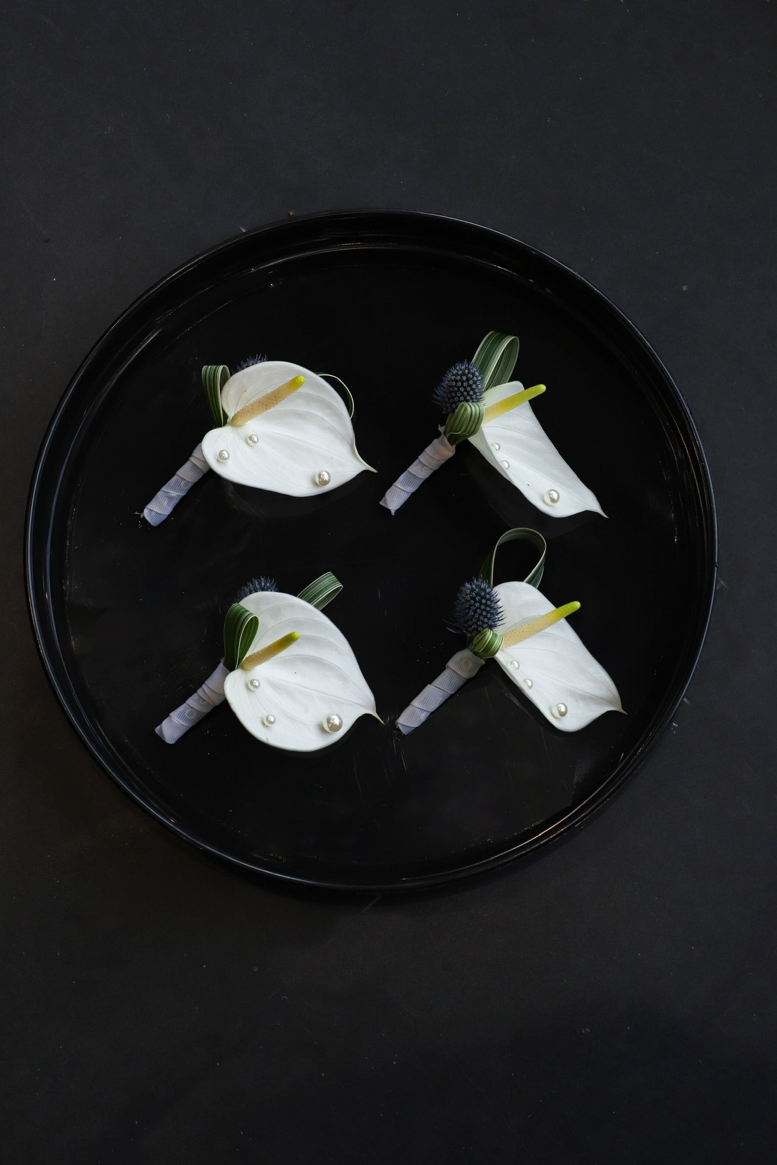 Four Pearl & Bloom corsages arranged on a black tray featuring white anthuriums, blue globe thistles, foliage and pearl accents tied with Just Bloom grey ribbon — Just Bloom Hong Kong