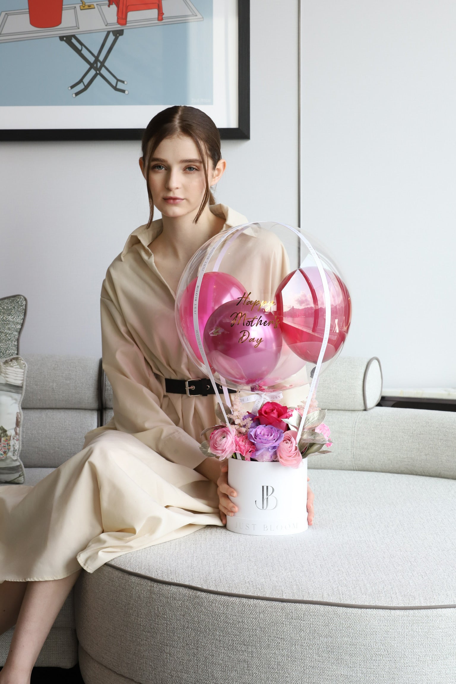 A model seated on a luxury sofa presenting Pink Reverie balloon flower box by Just Bloom Hong Kong, showcasing deep pink and rose blooms with a transparent bubble balloon in pink tones