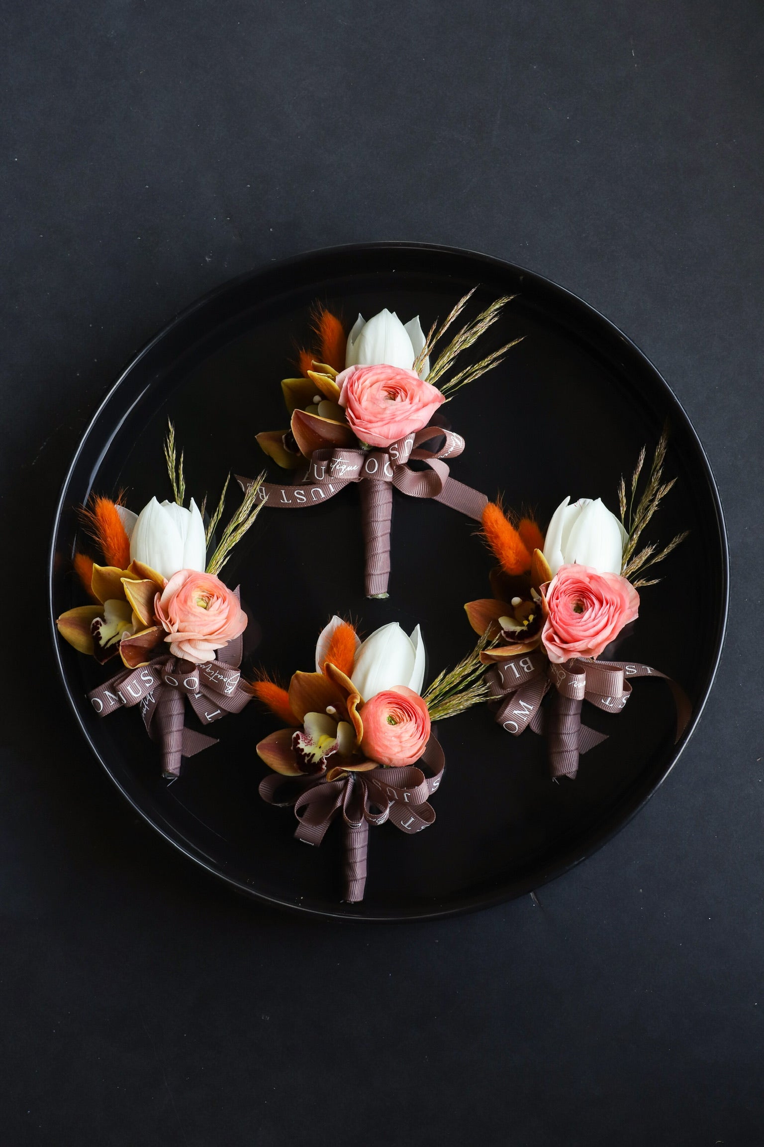 Four Rooted in Love corsages arranged on a black tray featuring white tulips, coral ranunculus, cymbidium orchids, dried lagurus ovatus and foliage tied with Just Bloom dark ribbon — Just Bloom Hong Kong