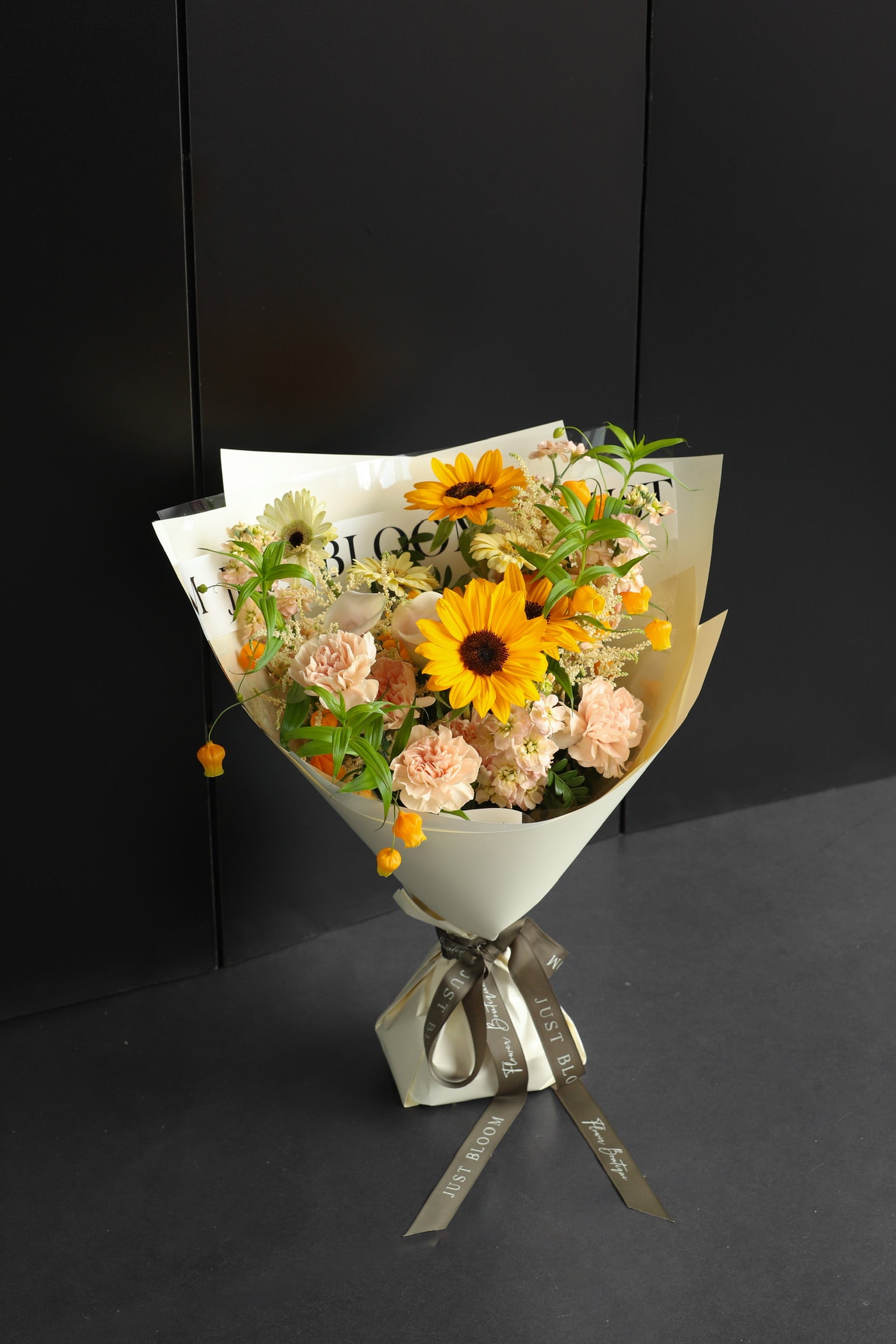Warm-toned fresh flower bouquet with sunflowers, blush carnations, and mixed seasonal blooms wrapped in white Just Bloom paper with grey ribbon against a dark background — Just Bloom Hong Kong