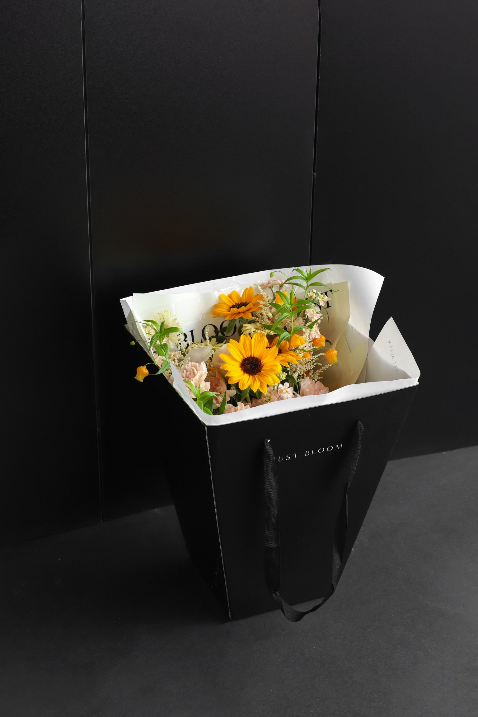 Just Bloom signature black gift bag carrying a fresh flower bouquet with sunflowers and mixed warm-toned blooms against a dark background — Just Bloom Hong Kong