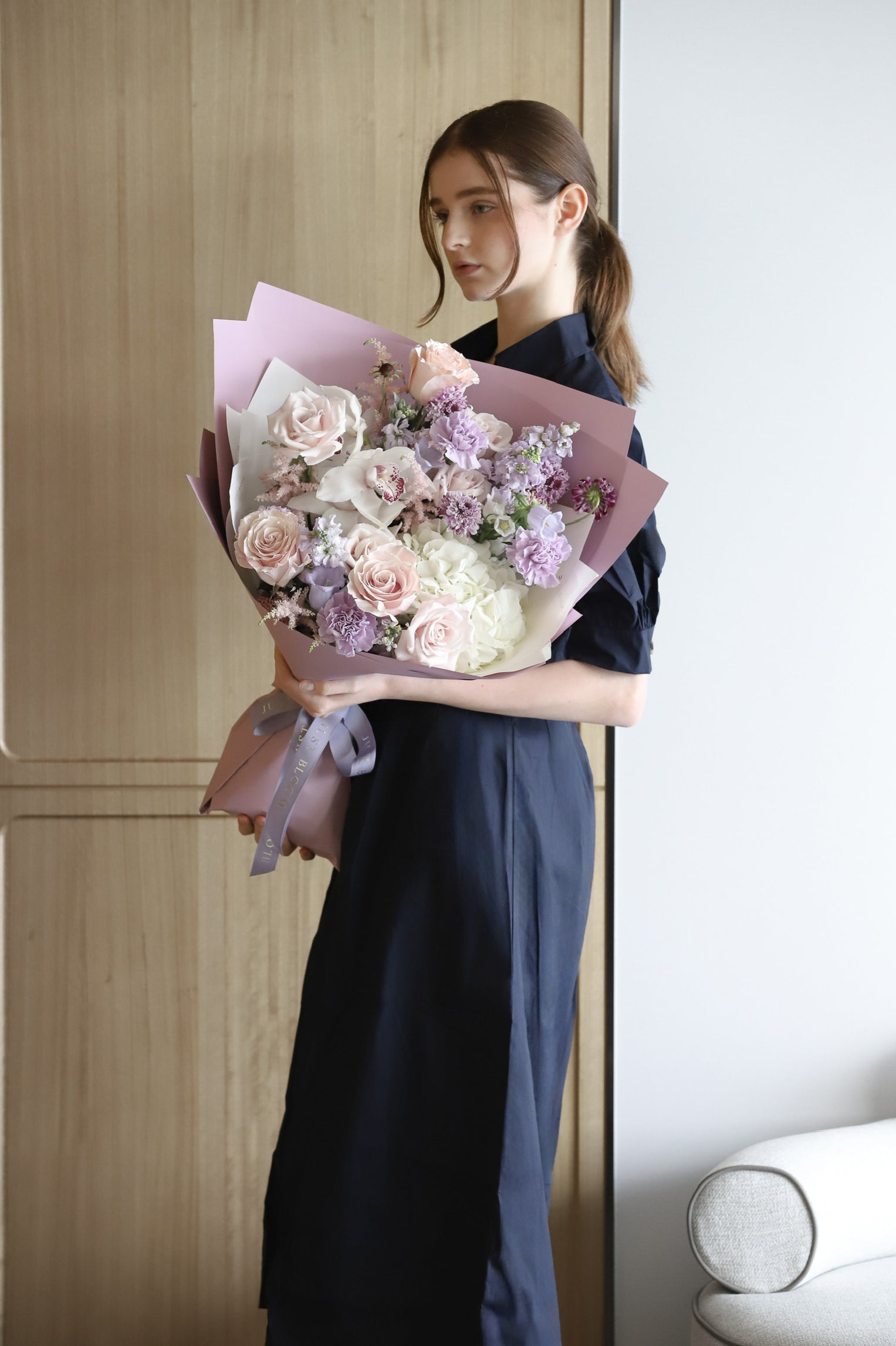 A model in a navy dress holding Soft & Timeless, a luxury bouquet in soft blush and lavender with dusty mauve wrapping and lilac ribbon — Just Bloom Hong Kong