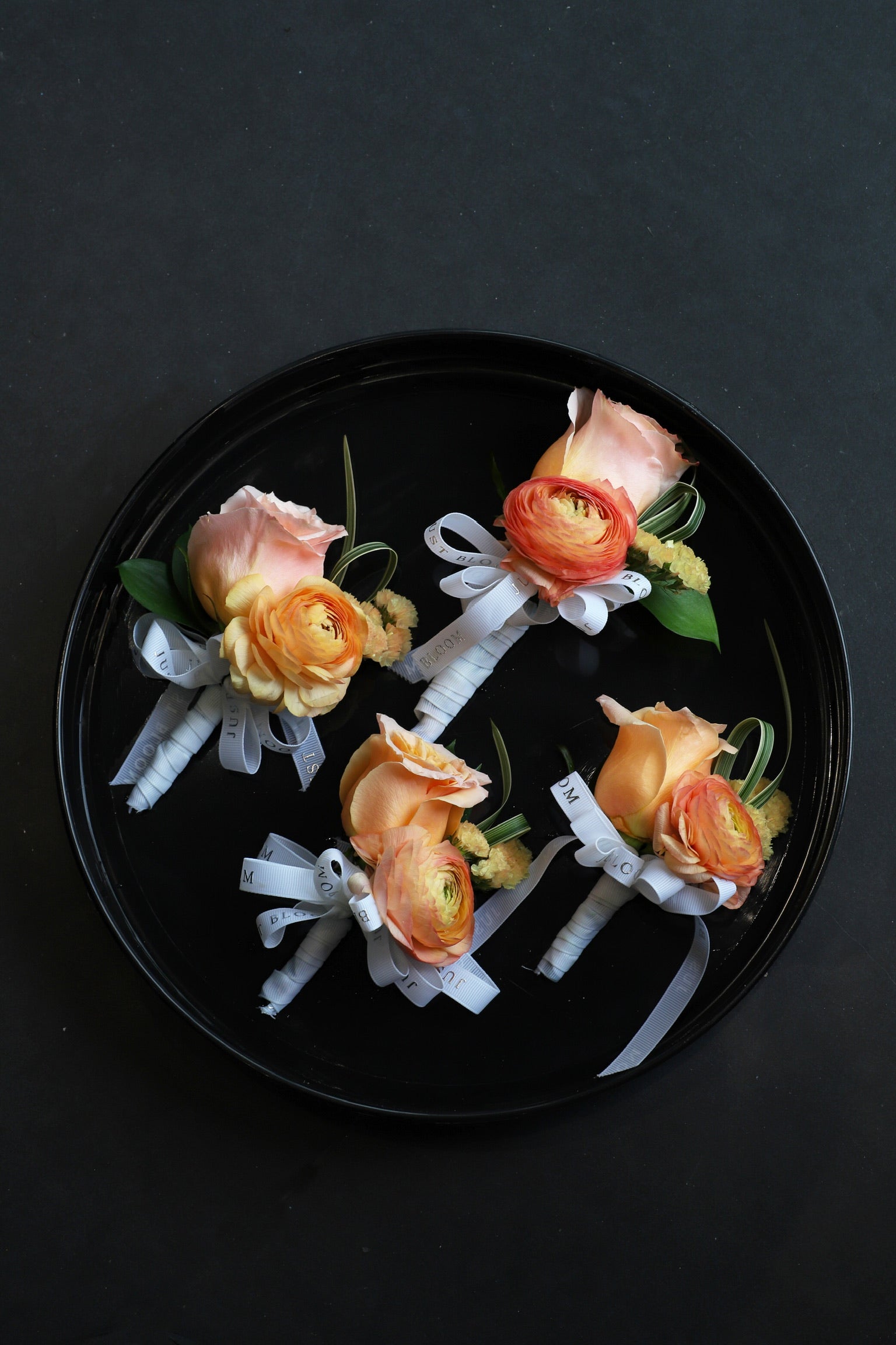 Four Sun Kissed corsages arranged on a black tray featuring peach Ecuadorian roses, orange ranunculus and myosotis tied with Just Bloom white ribbon — Just Bloom Hong Kong