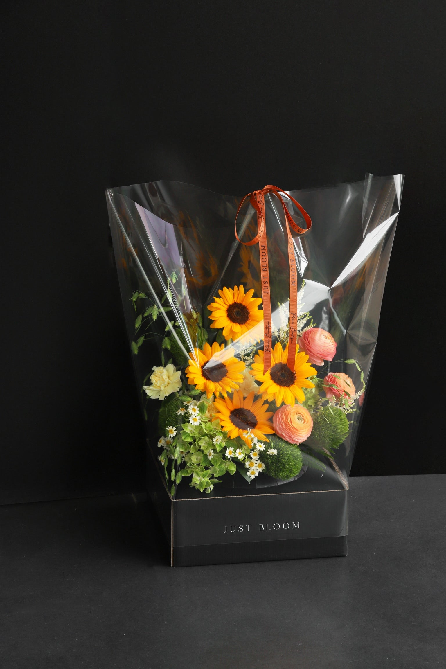 Just Bloom flower box arrangement in clear gift packaging with orange ribbon, showing warm-toned sunflowers and mixed seasonal blooms against a dark background — Just Bloom Hong Kong
