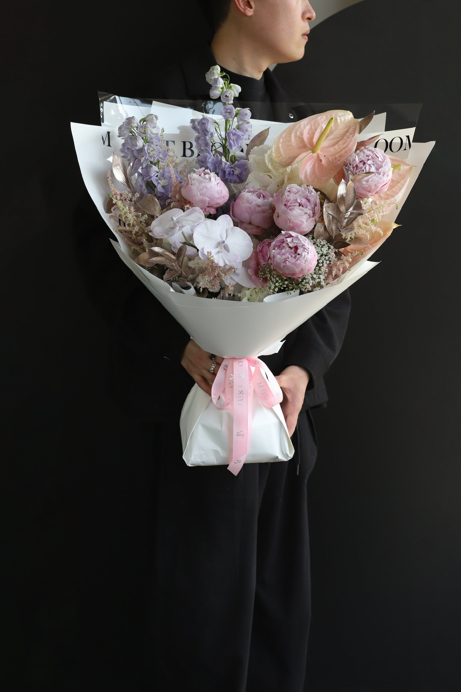 A model in black holding The Dreamer luxury bouquet by Just Bloom Hong Kong, showcasing pink peonies, lavender delphiniums, white orchids and anthuriums in white wrapping with pink ribbon
