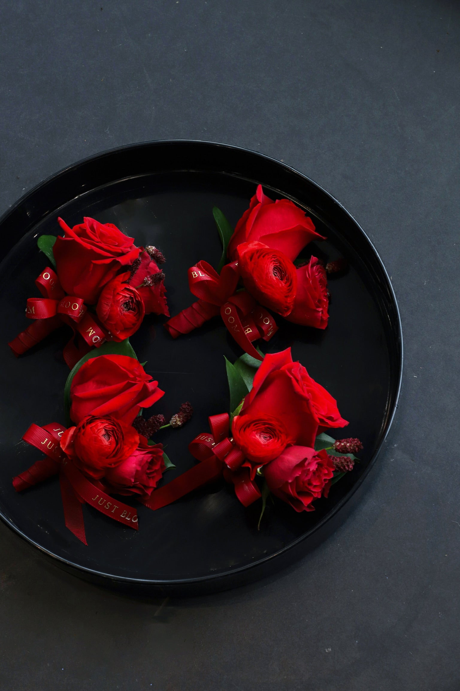 Close-up of four True Red wedding corsages on a black tray by Just Bloom Hong Kong, showcasing deep red roses, ranunculus and seasonal berries with Just Bloom deep red ribbon