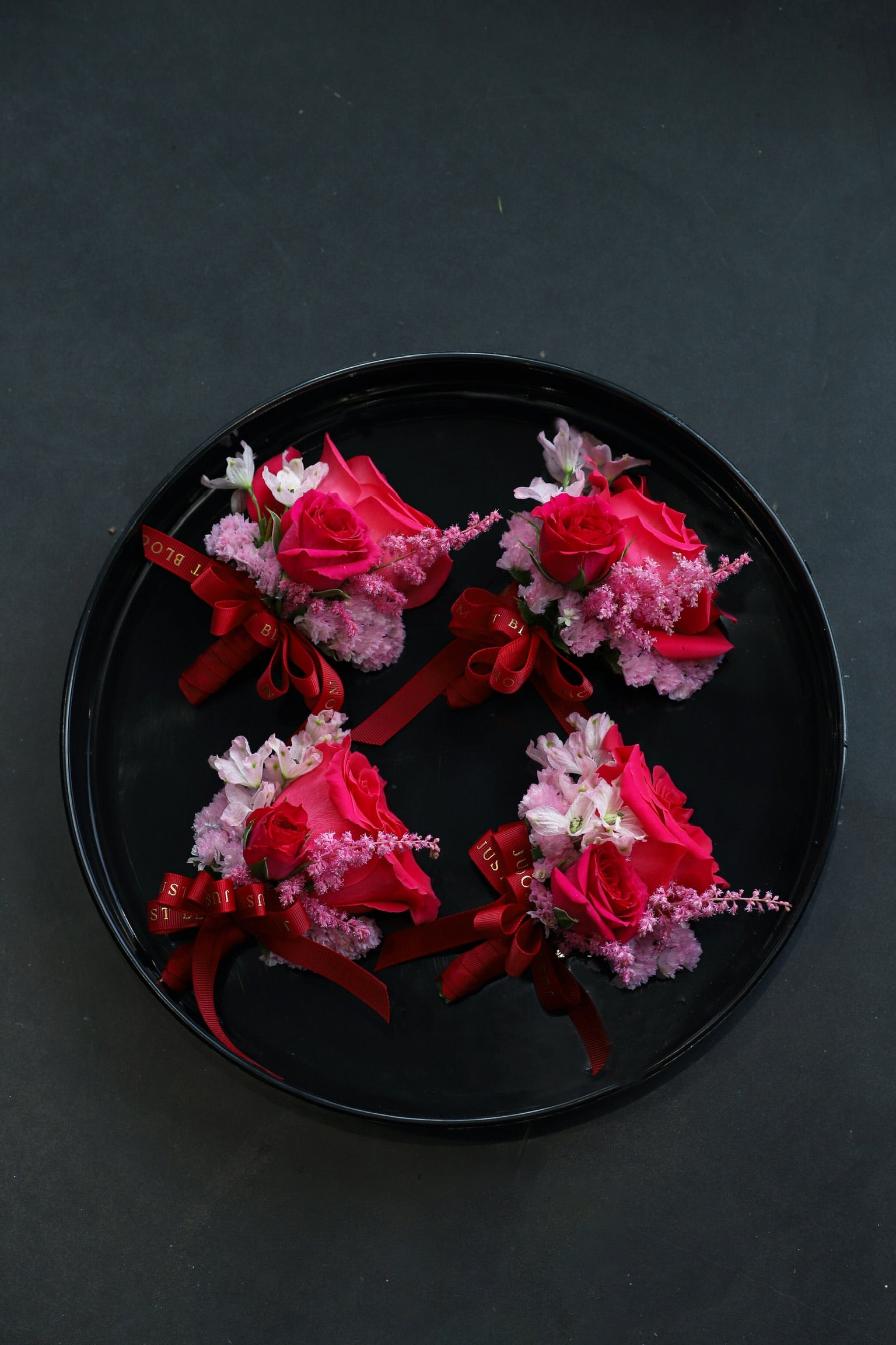Four Boutonnieres arranged on a black tray featuring fuchsia Ecuadorian roses, deep pink astilbes, sweet peas and myosotis tied with Just Bloom deep red ribbon — Just Bloom Hong Kong