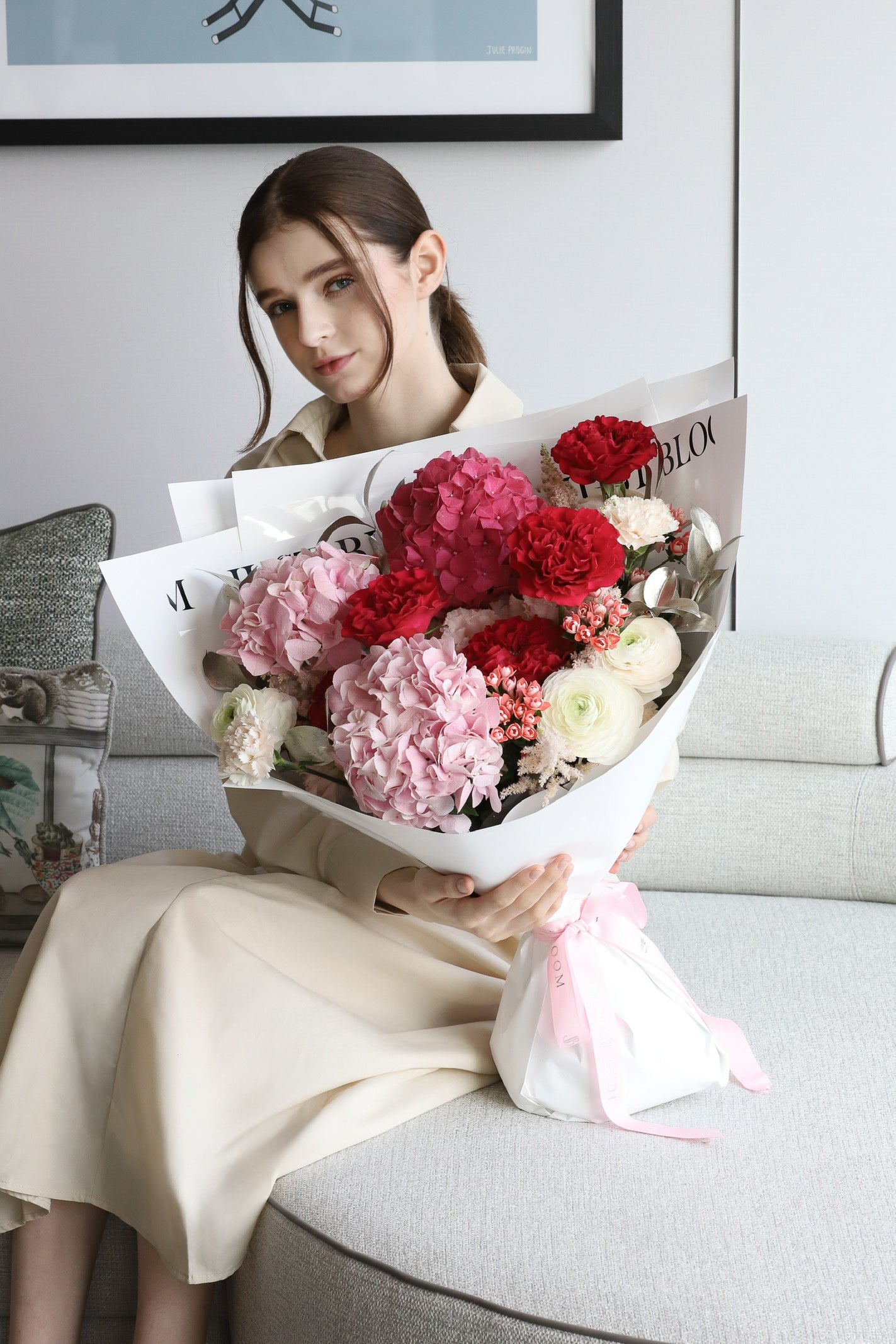 A model seated on a luxury sofa holding Wholehearted, a generous bouquet in vibrant pink, deep red and ivory with white wrapping and blush pink ribbon — Just Bloom Hong Kong