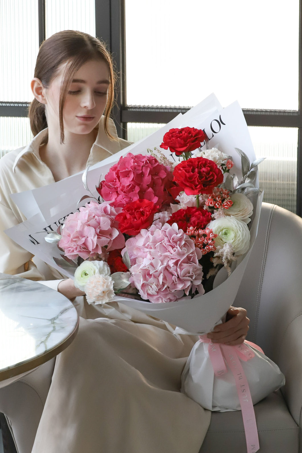 A model seated in a luxury chair looking down at Wholehearted bouquet by Just Bloom Hong Kong, showcasing voluminous pink hydrangeas, deep red carnations and ivory blooms in white wrapping
